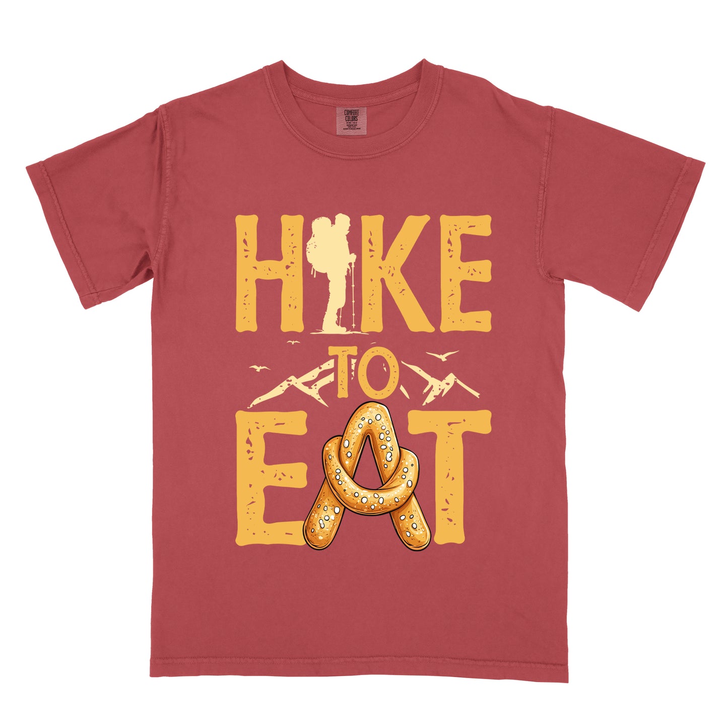 Crimson (Dark Pink) t-shirt with golden yellow distressed text reading "HIKE TO EAT". "HIKE" appears in large golden yellow letters at the top with a hiker silhouette integrated into the letter "I", "TO" in golden yellow text with mountain outlines on either side in the middle, and "EAT" in large golden yellow letters at the bottom with a pretzel shaped in an "A" replacing the letter "A". Small flying birds appear near "TO".