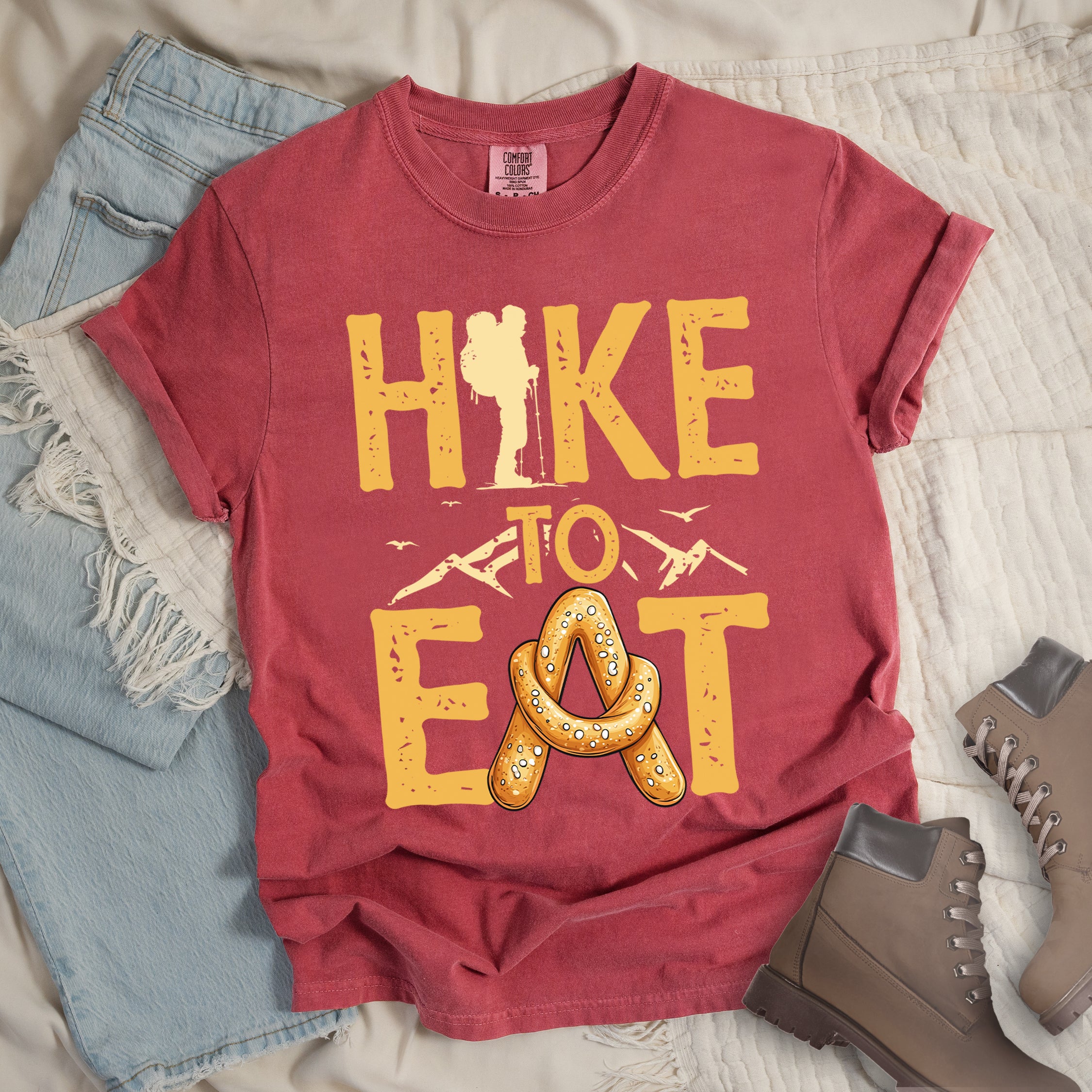 Crimson (Dark Pink) t-shirt with text reading "HIKE TO EAT". "HIKE" appears in golden yellow letters at the top with a hiker silhouette integrated into the letter "I", "TO" in golden yellow text with mountain outlines on either side in the middle, and "EAT" in large golden yellow letters at the bottom with a pretzel shaped in an "A" replacing the letter "A". Small flying birds appear near "TO". Features a classic crew neck and casual rolled sleeves, styled alongside light wash denim and brown hiking boots