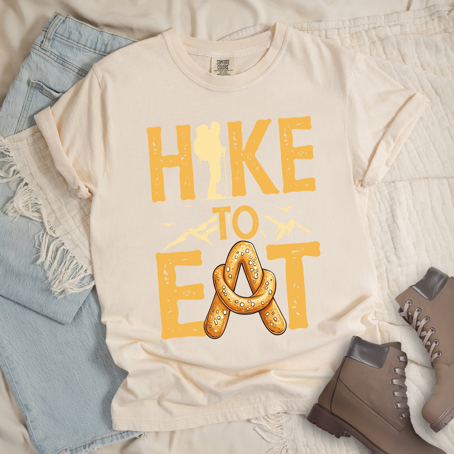 Ivory (Cream White) t-shirt with text reading "HIKE TO EAT". "HIKE" appears in golden yellow letters at the top with a hiker silhouette integrated into the letter "I", "TO" in golden yellow text with mountain outlines on either side in the middle, and "EAT" in large golden yellow letters at the bottom with a pretzel shaped in an "A" replacing the letter "A". Small flying birds appear near "TO". Features a classic crew neck and casual rolled sleeves, styled alongside light wash denim and brown hiking boots.