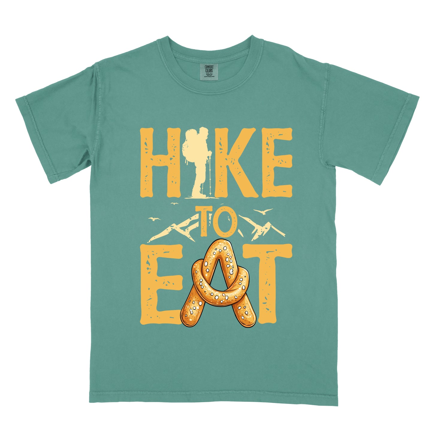Light Green t-shirt with golden yellow distressed text reading "HIKE TO EAT". "HIKE" appears in large golden yellow letters at the top with a hiker silhouette integrated into the letter "I", "TO" in golden yellow text with mountain outlines on either side in the middle, and "EAT" in large golden yellow letters at the bottom with a pretzel shaped in an "A" replacing the letter "A". Small flying birds appear near "TO".