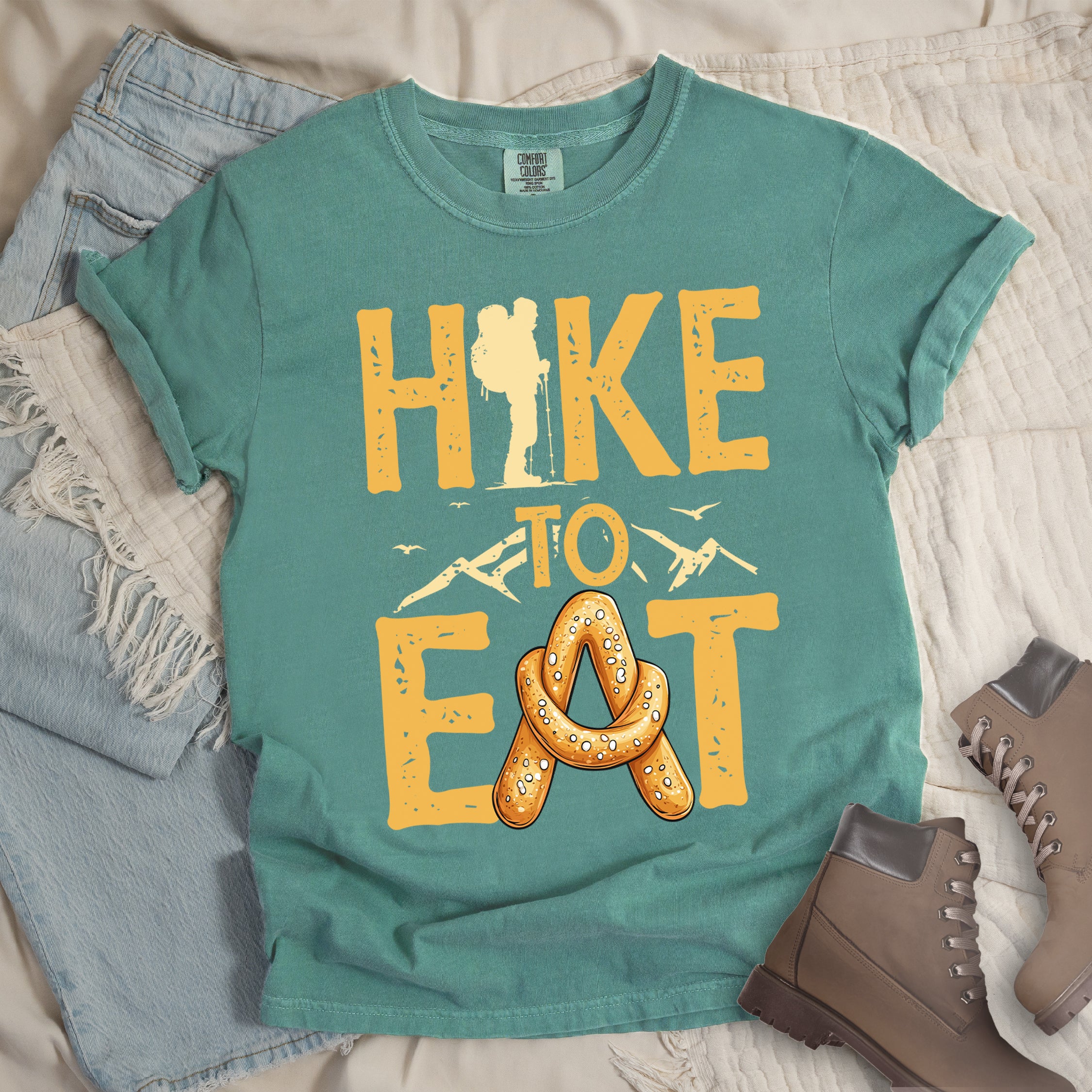 Light Green t-shirt with text reading "HIKE TO EAT". "HIKE" appears in golden yellow letters at the top with a hiker silhouette integrated into the letter "I", "TO" in golden yellow text with mountain outlines on either side in the middle, and "EAT" in large golden yellow letters at the bottom with a pretzel shaped in an "A" replacing the letter "A". Small flying birds appear near "TO". Features a classic crew neck and casual rolled sleeves, styled alongside light wash denim and brown hiking boots.
