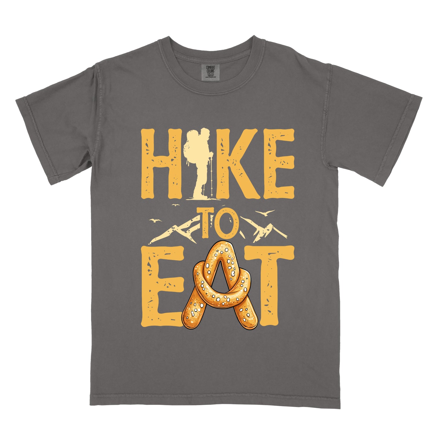 Pepper (Dark Gray) t-shirt with golden yellow distressed text reading "HIKE TO EAT". "HIKE" appears in large golden yellow letters at the top with a hiker silhouette integrated into the letter "I", "TO" in golden yellow text with mountain outlines on either side in the middle, and "EAT" in large golden yellow letters at the bottom with a pretzel shaped in an "A" replacing the letter "A". Small flying birds appear near "TO".