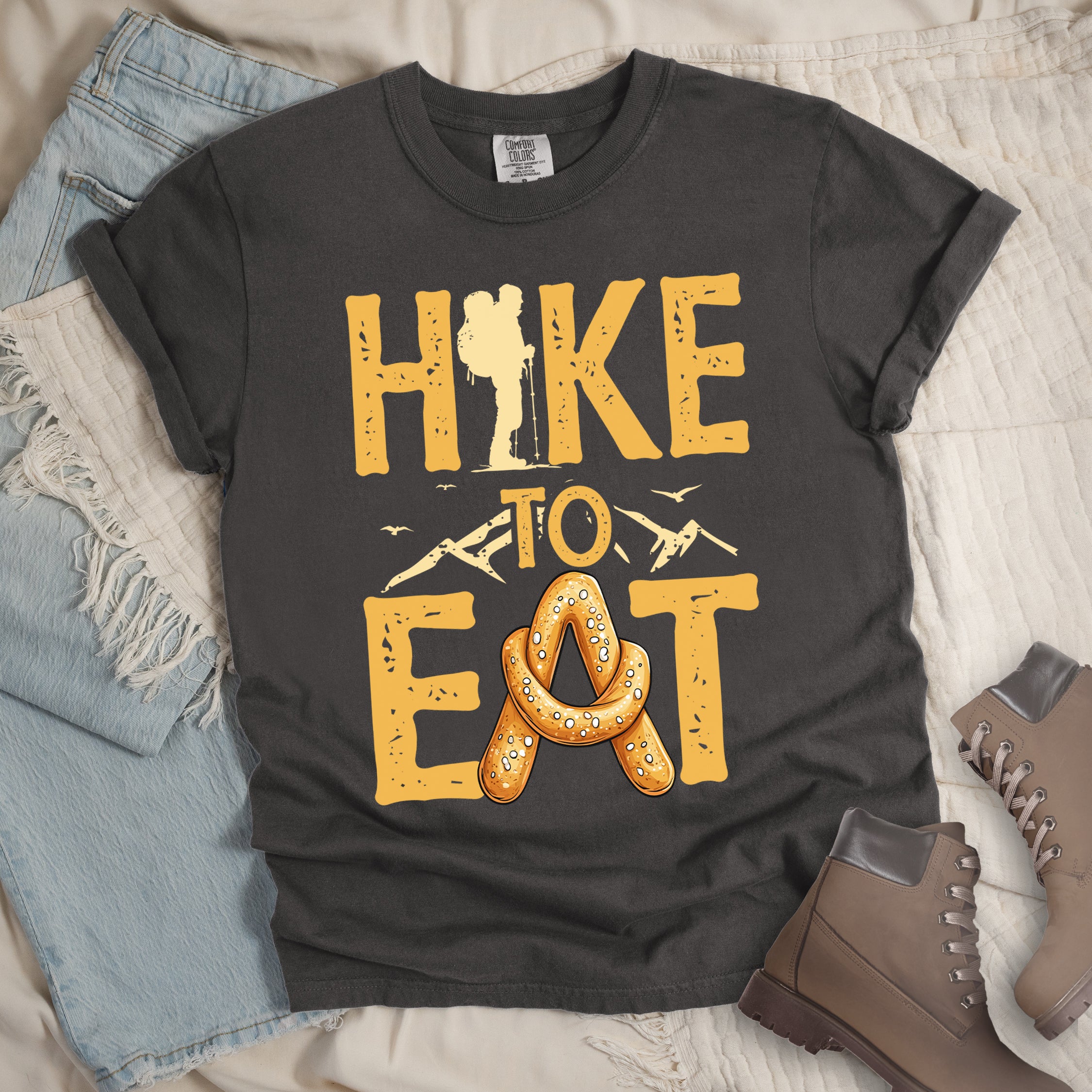 Pepper (Dark Gray) t-shirt with text reading "HIKE TO EAT". "HIKE" appears in golden yellow letters at the top with a hiker silhouette integrated into the letter "I", "TO" in golden yellow text with mountain outlines on either side in the middle, and "EAT" in large golden yellow letters at the bottom with a pretzel shaped in an "A" replacing the letter "A". Small flying birds appear near "TO". Features a classic crew neck and casual rolled sleeves, styled alongside light wash denim and brown hiking boots.