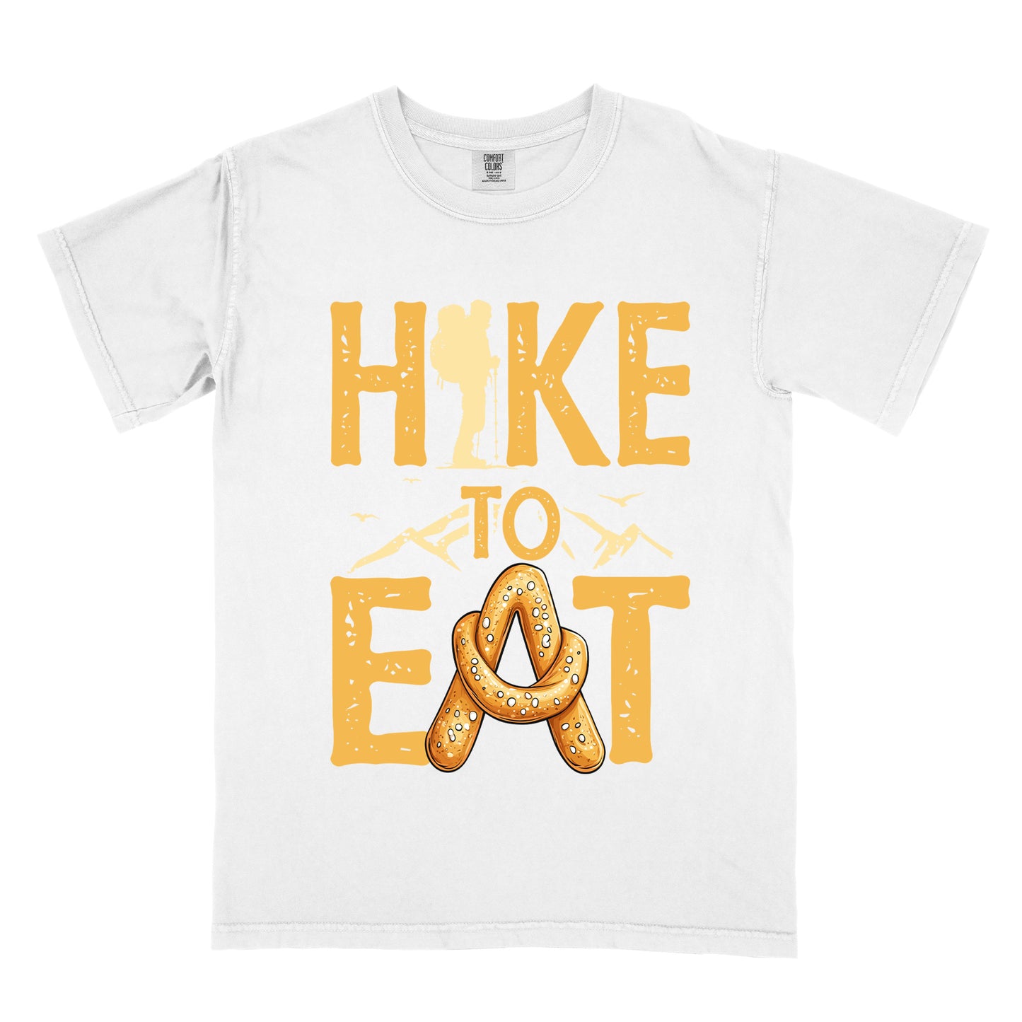 White t-shirt with golden yellow distressed text reading "HIKE TO EAT". "HIKE" appears in large golden yellow letters at the top with a hiker silhouette integrated into the letter "I", "TO" in golden yellow text with mountain outlines on either side in the middle, and "EAT" in large golden yellow letters at the bottom with a pretzel shaped in an "A" replacing the letter "A". Small flying birds appear near "TO".