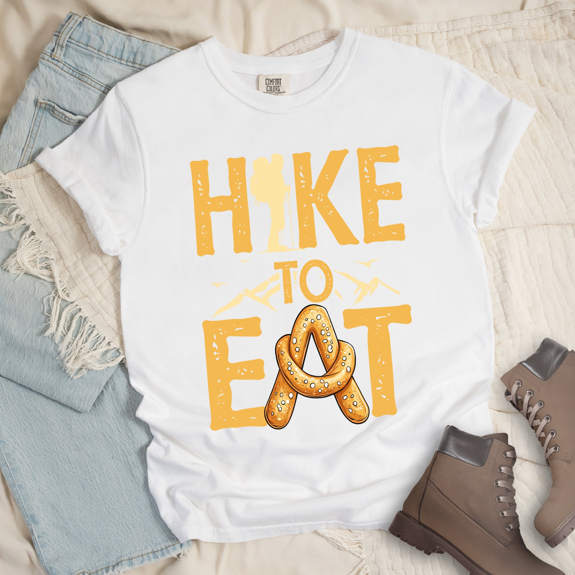 White t-shirt with text reading "HIKE TO EAT". "HIKE" appears in golden yellow letters at the top with a hiker silhouette integrated into the letter "I", "TO" in golden yellow text with mountain outlines on either side in the middle, and "EAT" in large golden yellow letters at the bottom with a pretzel shaped in an "A" replacing the letter "A". Small flying birds appear near "TO". Features a classic crew neck and casual rolled sleeves, styled alongside light wash denim and brown hiking boots.