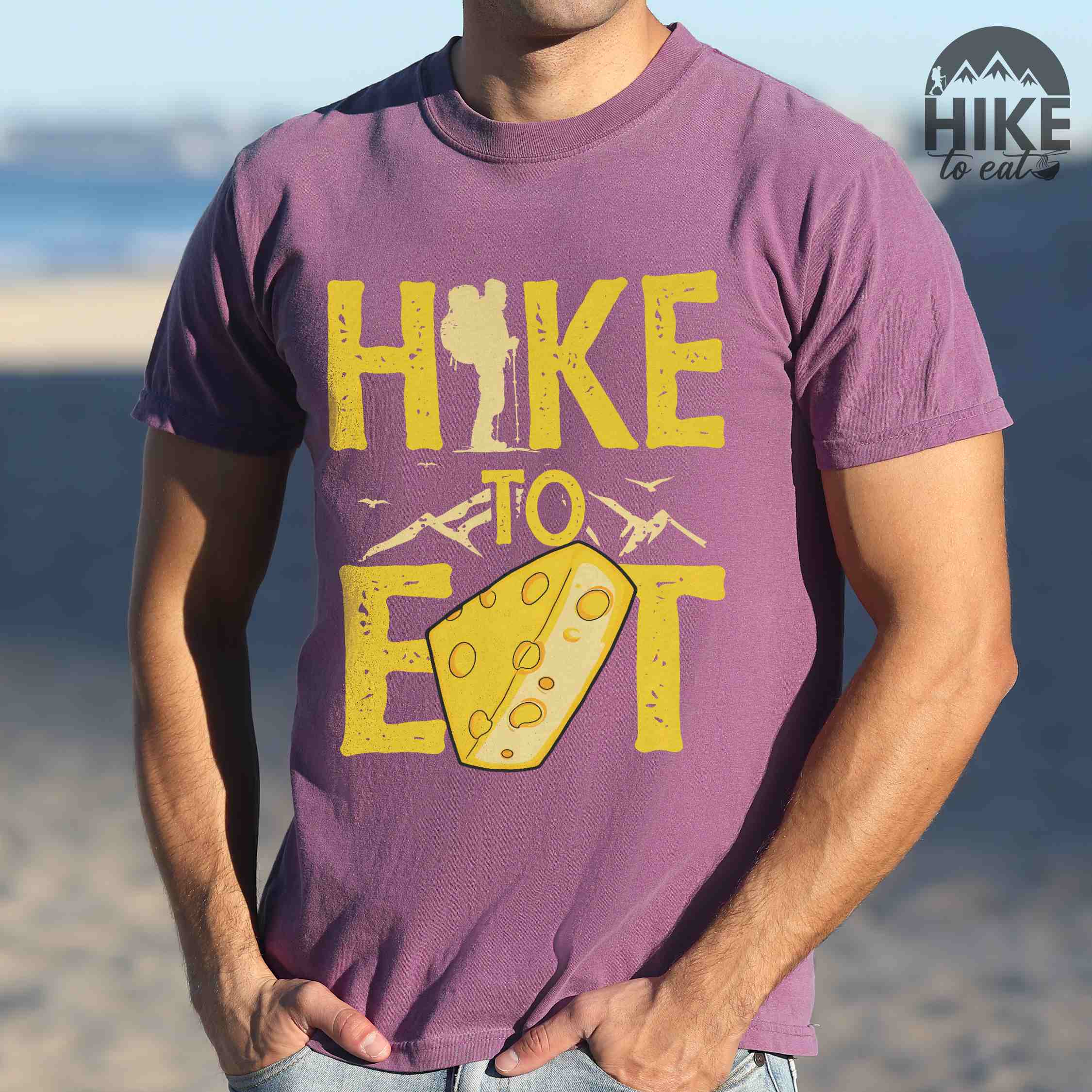 Man wearing berry "Hike to Eat" colored statement tee with bold typography, mountain, illustrated slice of cheese, and backpacker for cheese lovers.
