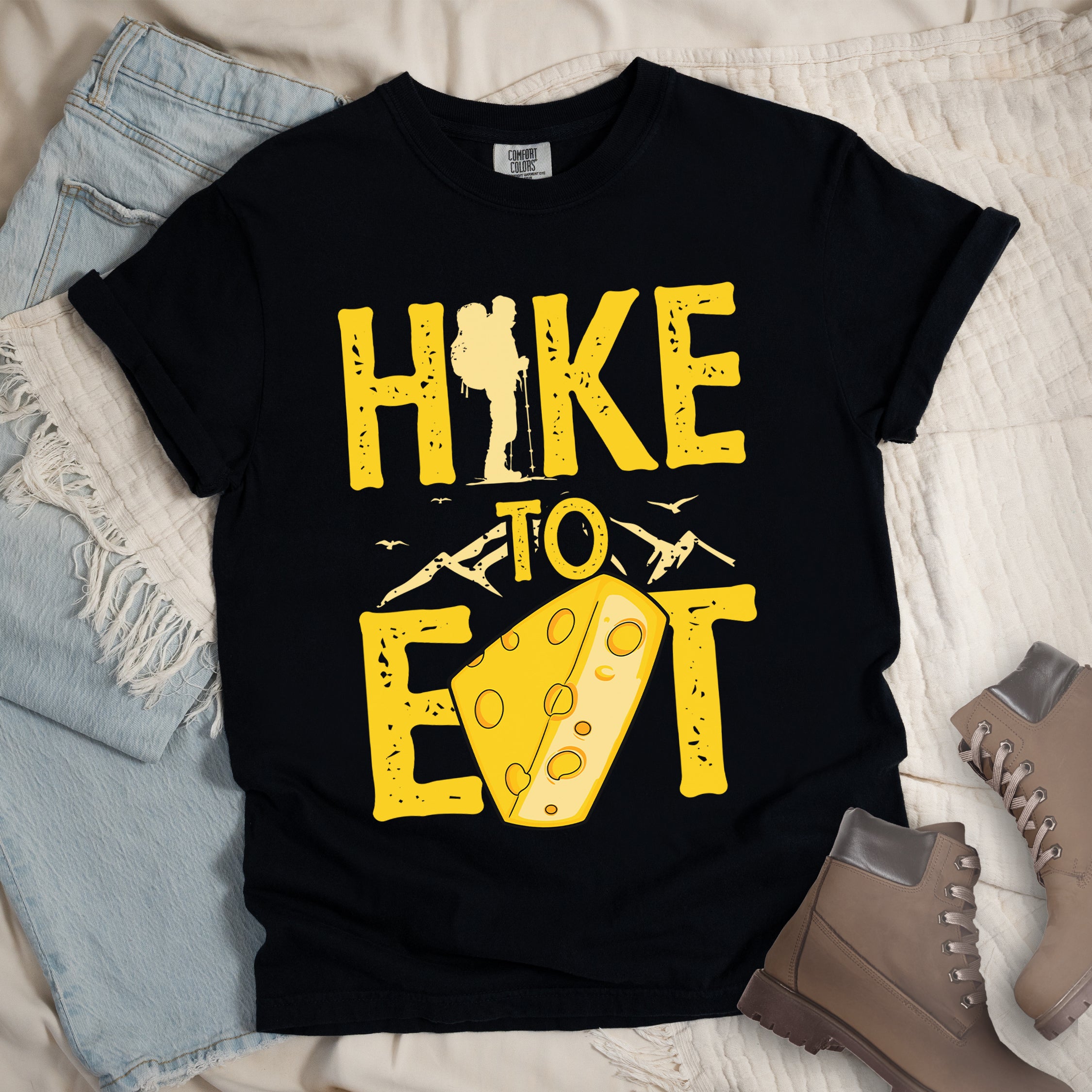 Black t-shirt with yellow distressed text reading "HIKE TO EAT". "HIKE" appears in large yellow letters at the top with a hiker silhouette as the letter "I", "TO" in yellow text with mountain outlines on either side in the middle, and "EAT" in large yellow letters at the bottom with a wedge of Swiss cheese positioned between "E" and "T". Small flying birds appear near "TO". The shirt features a classic crew neck and casual rolled sleeves, styled alongside light wash denim and brown hiking boots.