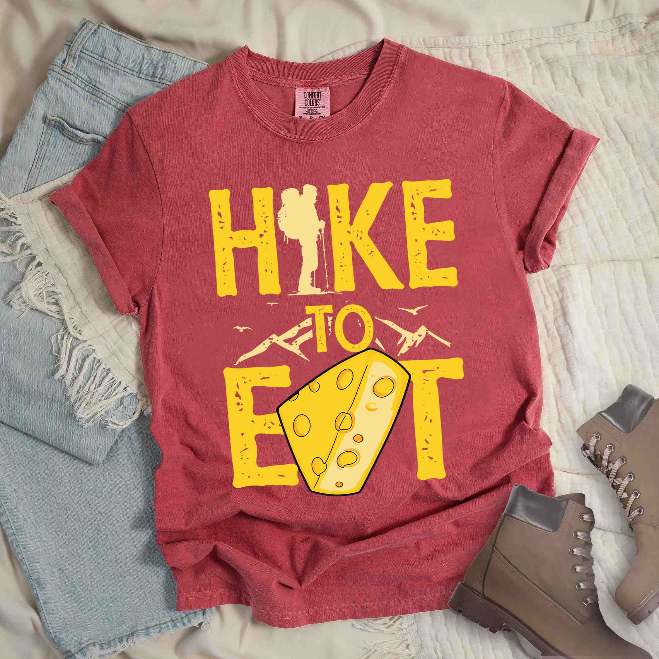 Crimson "Hike to Eat" colored statement tee with bold typography, mountain, illustrated slice of cheese, and backpacker for cheese lovers.