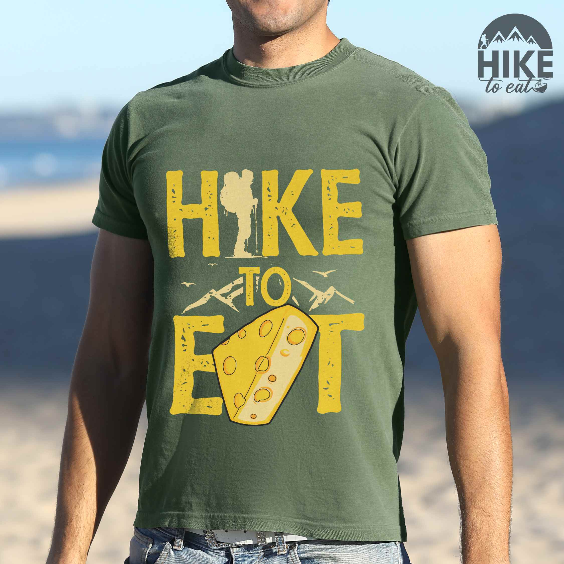 Man wearing moss "Hike to Eat" colored statement tee with bold typography, mountain, illustrated slice of cheese, and backpacker for cheese lovers.