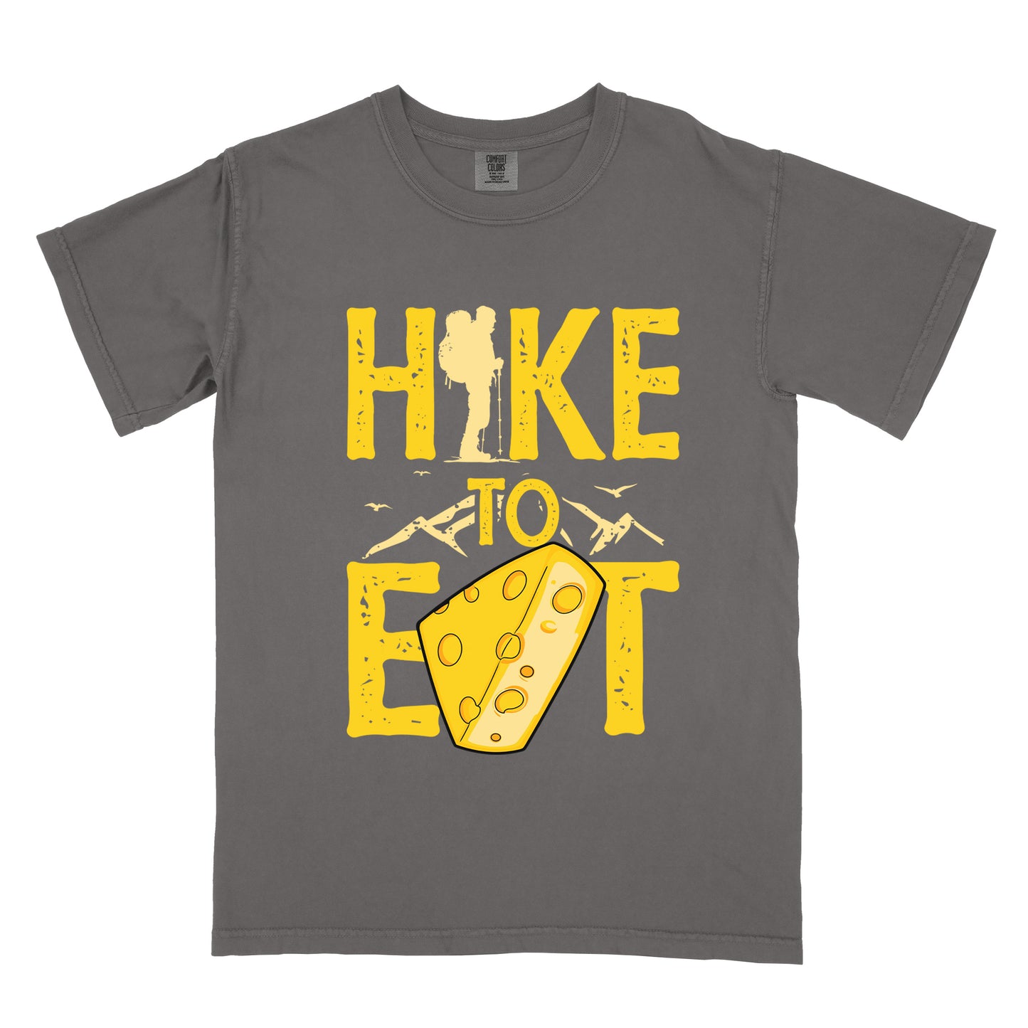Pepper (Dark Gray) t-shirt with yellow distressed text reading "HIKE TO EAT". "HIKE" appears in large yellow letters at the top with a hiker silhouette integrated into the letter "I", "TO" in yellow text with mountain outlines on either side in the middle, and "EAT" in large yellow letters at the bottom with a wedge of Swiss cheese positioned between "E" and "T". Small flying birds appear near "TO". The design connects hiking with eating cheese as motivation.