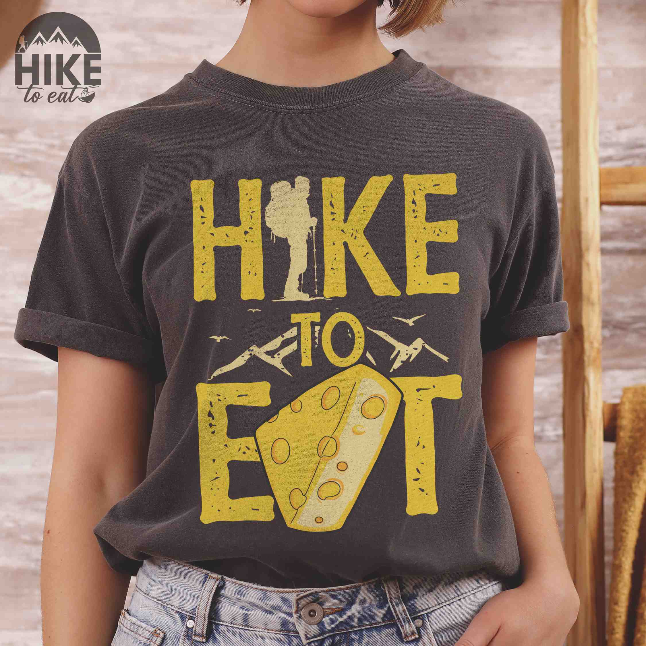 Woman wearing pepper "Hike to Eat" colored statement tee with bold typography, mountain, illustrated slice of cheese, and backpacker for cheese lovers.
