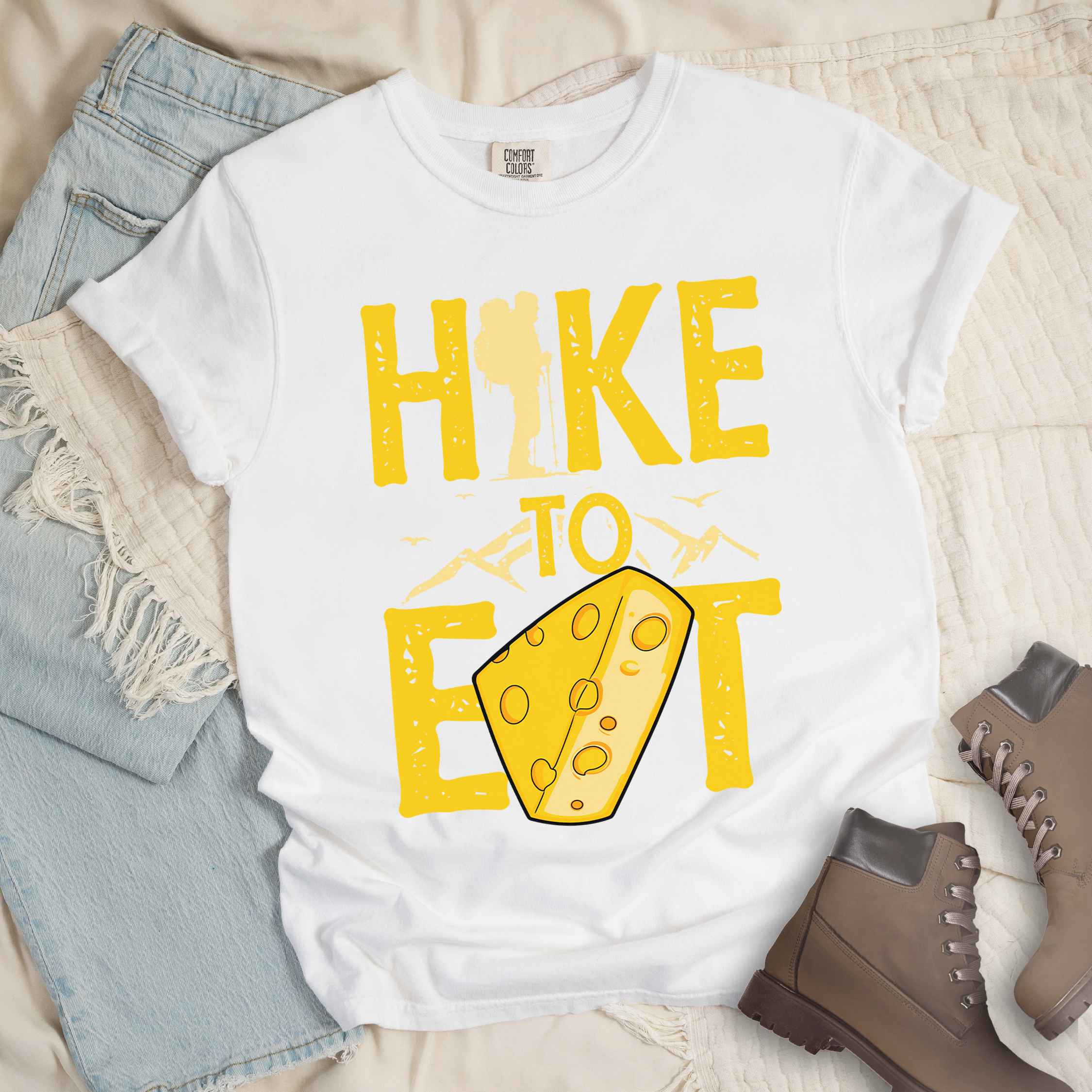 White "Hike to Eat" colored statement tee with bold typography, mountain, illustrated slice of cheese, and backpacker for cheese lovers.