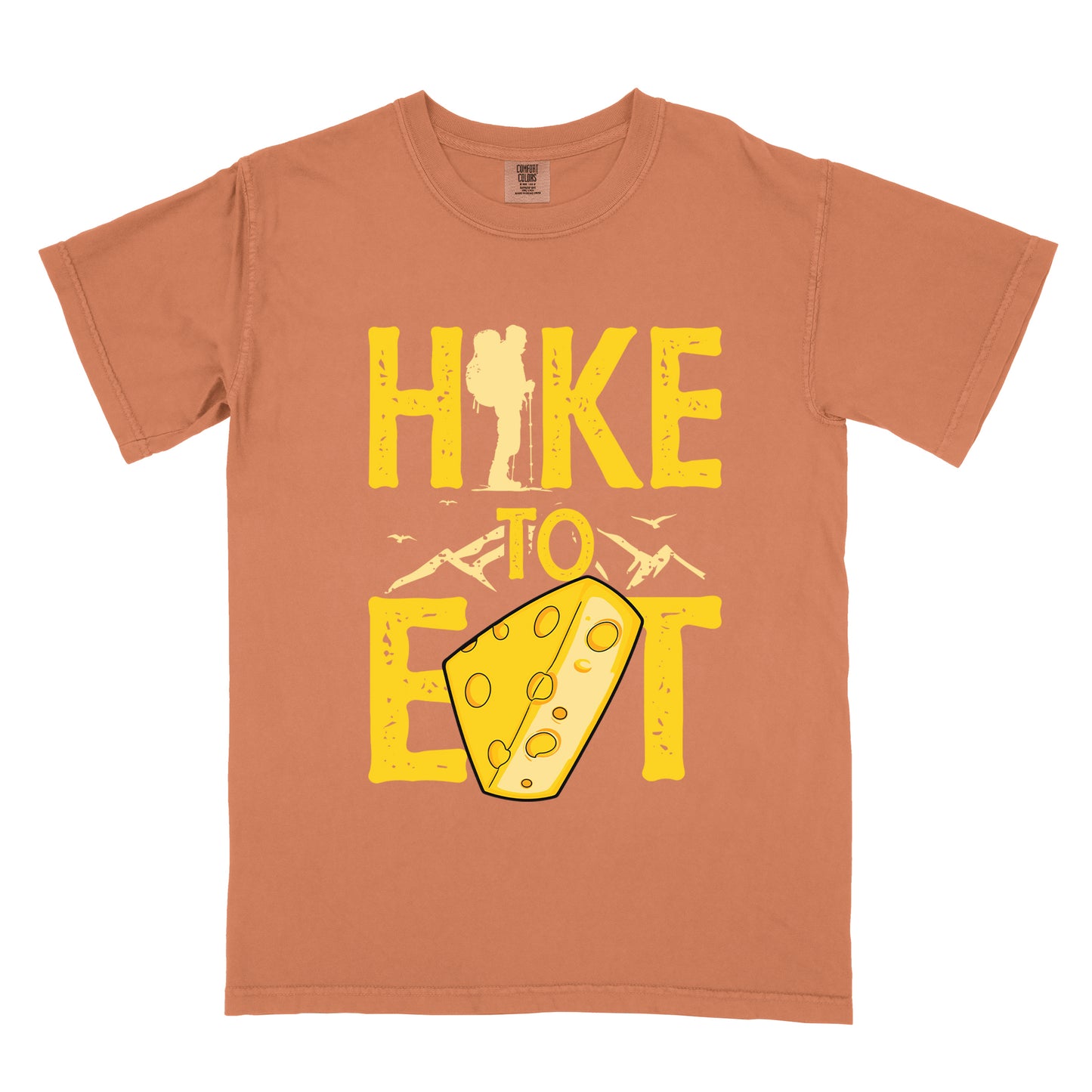 Yam (Brown) t-shirt with yellow distressed text reading "HIKE TO EAT". "HIKE" appears in large yellow letters at the top with a hiker silhouette integrated into the letter "I", "TO" in yellow text with mountain outlines on either side in the middle, and "EAT" in large yellow letters at the bottom with a wedge of Swiss cheese positioned between "E" and "T". Small flying birds appear near "TO". The design connects hiking with eating cheese as motivation.