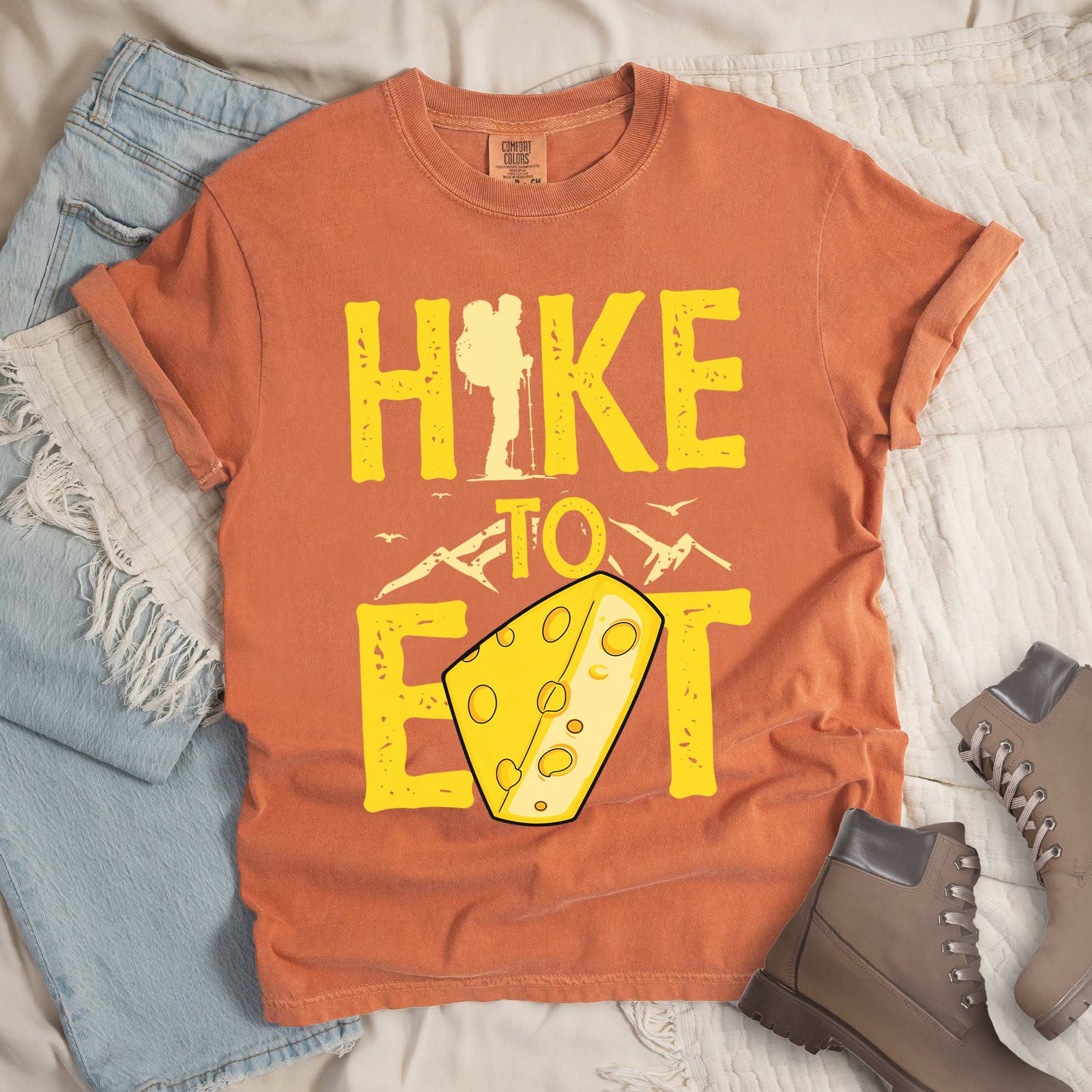 Yam (Brown) t-shirt with yellow distressed text reading "HIKE TO EAT". "HIKE" appears in large yellow letters at the top with a hiker silhouette integrated into the letter "I", "TO" in yellow text with mountain outlines on either side in the middle, and "EAT" in large yellow letters at the bottom with a wedge of Swiss cheese positioned between "E" and "T". Small flying birds appear near "TO". Features a classic crew neck and casual rolled sleeves, alongside light wash denim and brown hiking boots.