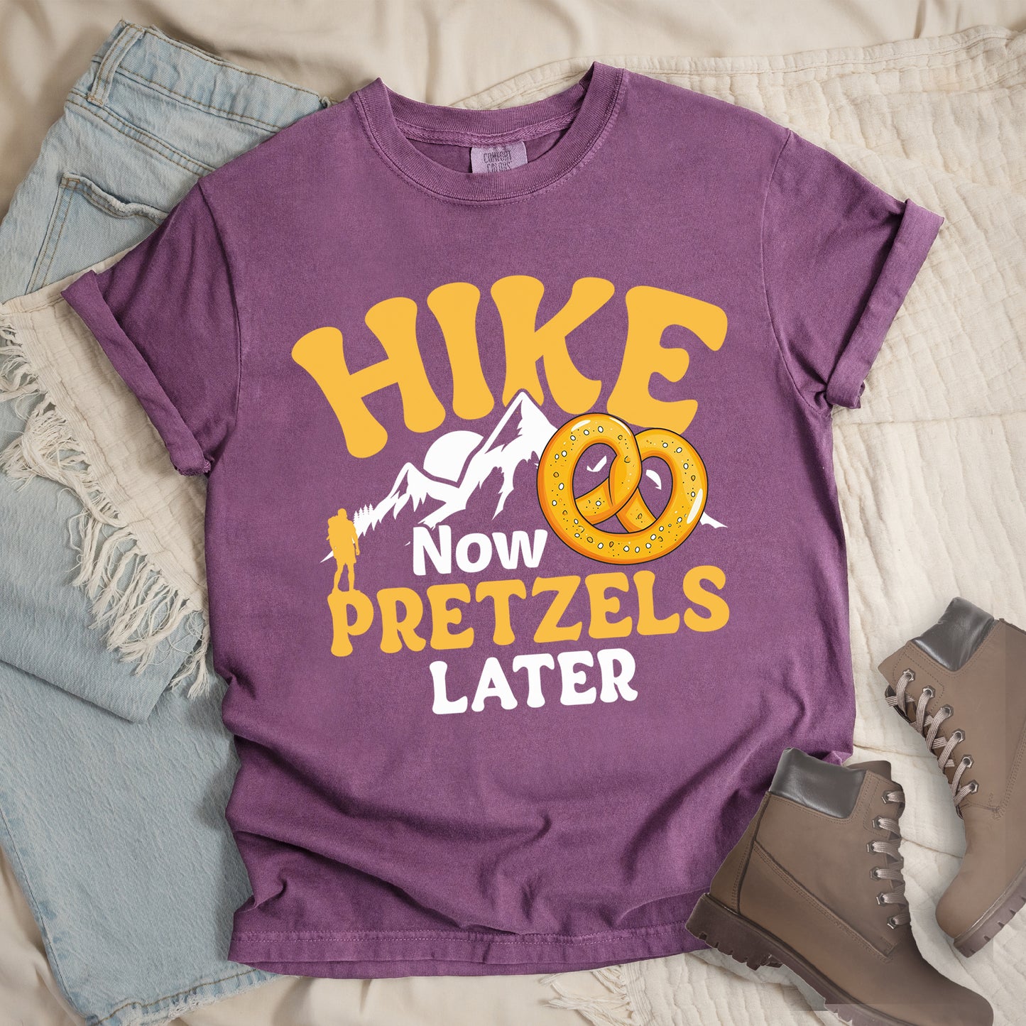 Berry (Dark Violet) t-shirt with "HIKE NOW PRETZELS LATER" text design. "HIKE" appears at the top in yellow letters, "NOW" in white, "PRETZELS" in yellow, and "LATER" in white. The design includes white mountain silhouettes with a small yellow hiker figure and an illustration of a golden pretzel with salt. Features a classic crew neck and casual rolled sleeves, styled alongside light wash denim and brown hiking boots.