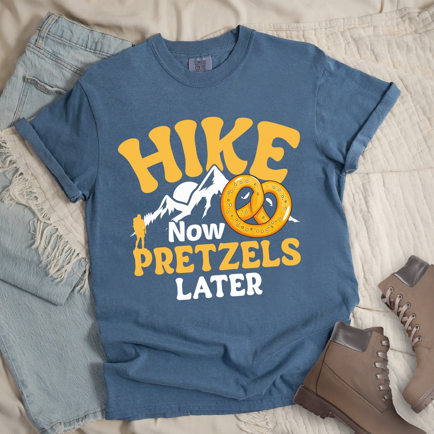 Blue Jean (Denim Blue) t-shirt with "HIKE NOW PRETZELS LATER" text design. "HIKE" appears at the top in yellow letters, "NOW" in white, "PRETZELS" in yellow, and "LATER" in white. The design includes white mountain silhouettes with a small yellow hiker figure and an illustration of a golden pretzel with salt. Features a classic crew neck and casual rolled sleeves, styled alongside light wash denim and brown hiking boots.