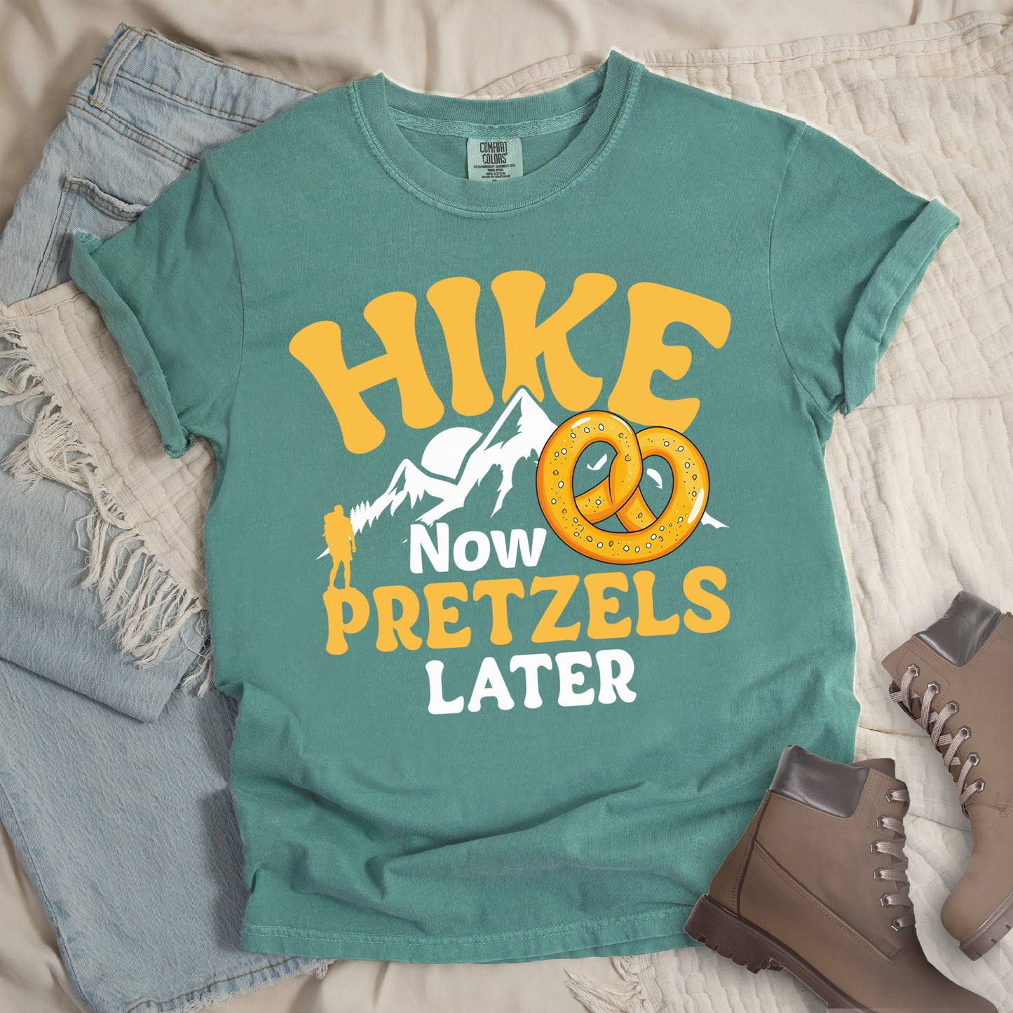 Light Green t-shirt with "HIKE NOW PRETZELS LATER" text design. "HIKE" appears at the top in yellow letters, "NOW" in white, "PRETZELS" in yellow, and "LATER" in white. The design includes white mountain silhouettes with a small yellow hiker figure and an illustration of a golden pretzel with salt. Features a classic crew neck and casual rolled sleeves, styled alongside light wash denim and brown hiking boots.