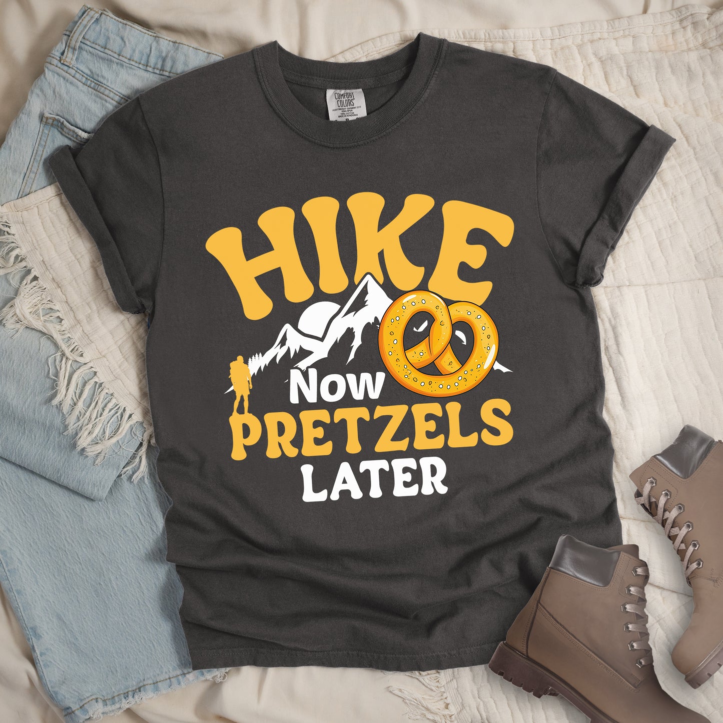 Pepper (Dark Gray) t-shirt with "HIKE NOW PRETZELS LATER" text design. "HIKE" appears at the top in yellow letters, "NOW" in white, "PRETZELS" in yellow, and "LATER" in white. The design includes white mountain silhouettes with a small yellow hiker figure and an illustration of a golden pretzel with salt. Features a classic crew neck and casual rolled sleeves, styled alongside light wash denim and brown hiking boots.