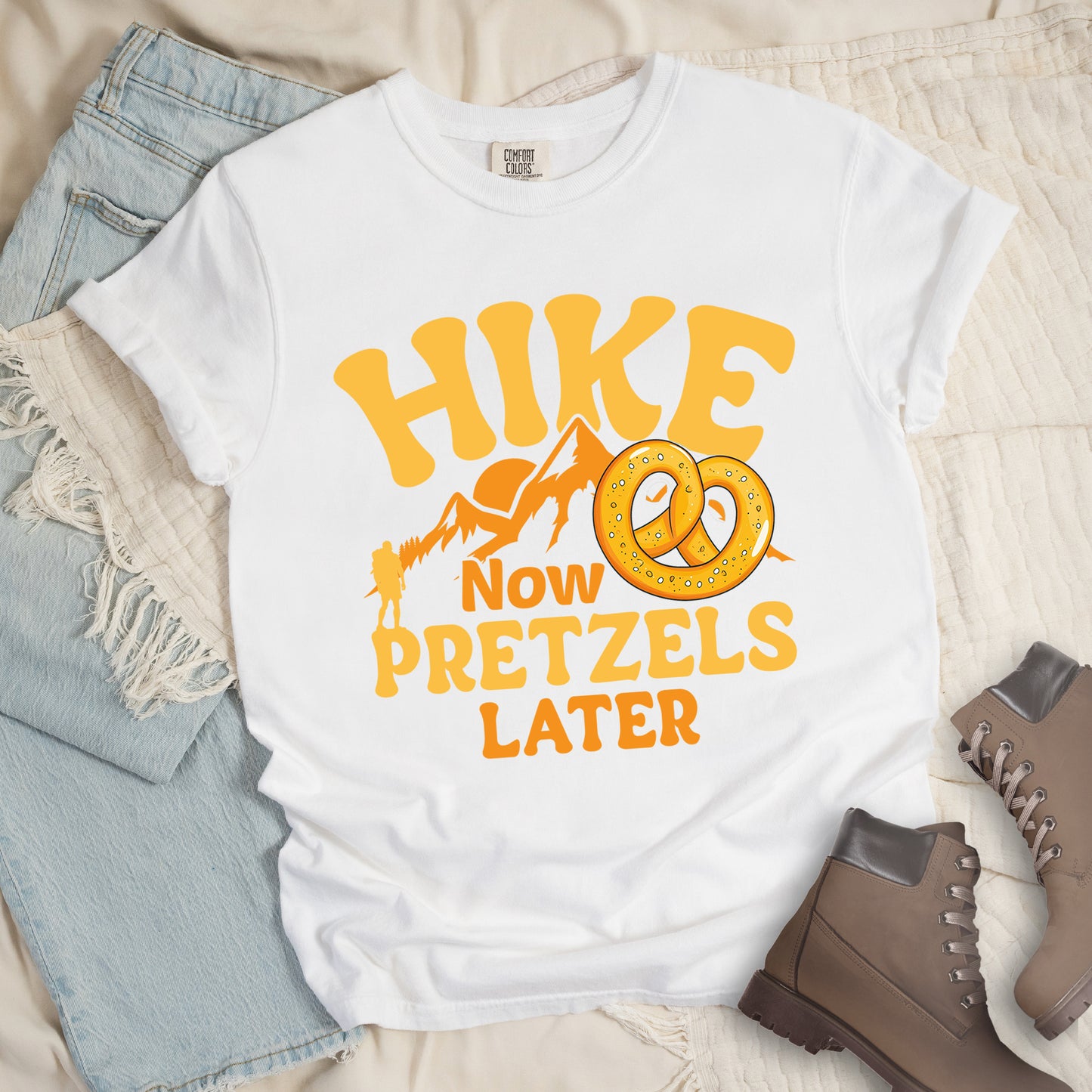 White t-shirt with "HIKE NOW PRETZELS LATER" text design. "HIKE" appears at the top in yellow letters, "NOW" in orange, "PRETZELS" in yellow, and "LATER" in orange. The design includes orange mountain silhouettes with a small yellow hiker figure and an illustration of a golden pretzel with salt. The playful text arrangement emphasizes hiking first and enjoying pretzels as a reward afterward. Features a classic crew neck and casual rolled sleeves, styled alongside light wash denim and brown hiking boots.