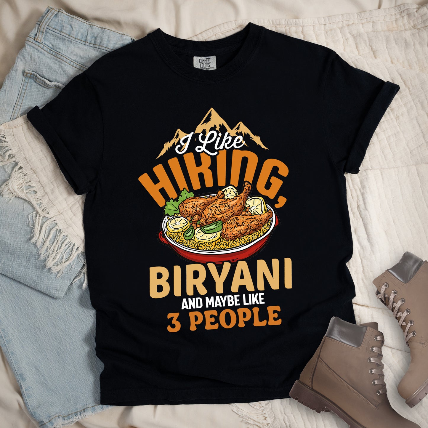 Black t-shirt with text reading "I Like HIKING, BIRYANI AND MAYBE LIKE 3 PEOPLE". Gold mountain silhouettes appear above "I Like" (in white script). "HIKING" is in large orange letters, below which sits a detailed illustration of a red bowl filled with biryani rice and chicken. "BIRYANI" appears in gold letters, while "AND MAYBE LIKE" is in small white text and "3 PEOPLE" in orange. Features a classic crew neck and casual rolled sleeves, styled alongside light wash denim and brown hiking boots.
