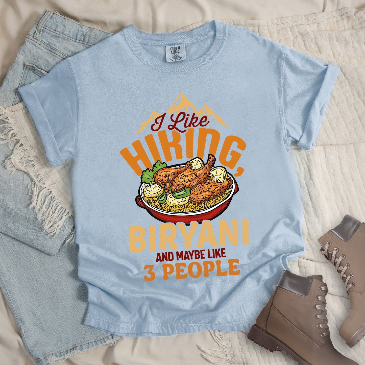Chambray (Light Blue) t-shirt with text "I Like HIKING, BIRYANI AND MAYBE LIKE 3 PEOPLE". Gold mountain silhouettes appear above "I Like" (in dark brown script). "HIKING" is in large orange letters, below which sits a detailed illustration of a red bowl filled with biryani rice and chicken. "BIRYANI" appears in gold letters, while "AND MAYBE LIKE" is in small dark brown text and "3 PEOPLE" in orange. Features a classic crew neck and casual rolled sleeves, alongside light wash denim and brown hiking boots.