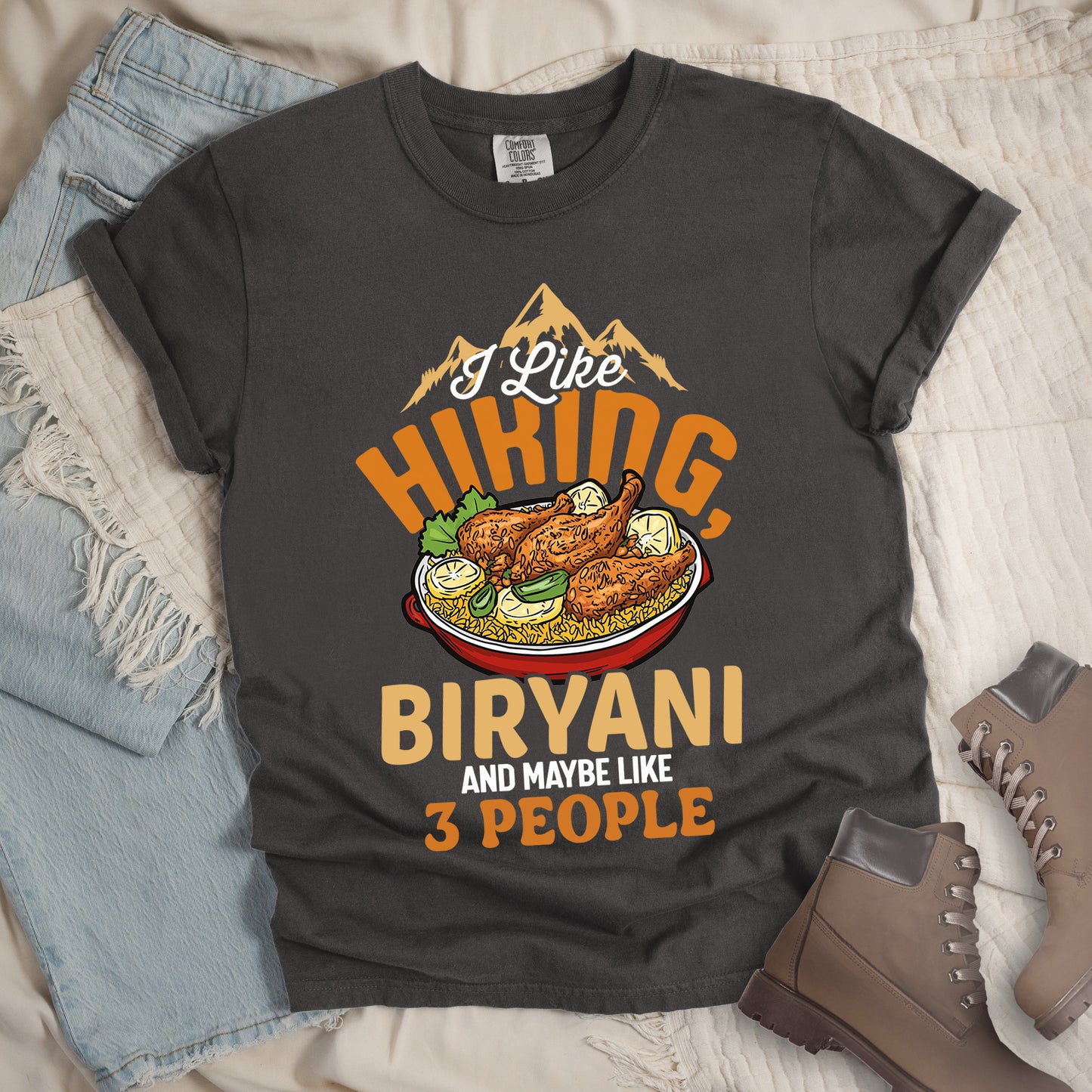 Pepper (Dark Gray) t-shirt with text reading "I Like HIKING, BIRYANI AND MAYBE LIKE 3 PEOPLE". Gold mountain silhouettes appear above "I Like" (in white script). "HIKING" is in large orange letters, below which sits a detailed illustration of a red bowl filled with biryani rice and chicken. "BIRYANI" appears in gold letters, while "AND MAYBE LIKE" is in small white text and "3 PEOPLE" in orange. Features a classic crew neck and casual rolled sleeves, styled alongside light wash denim and brown hiking boots.
