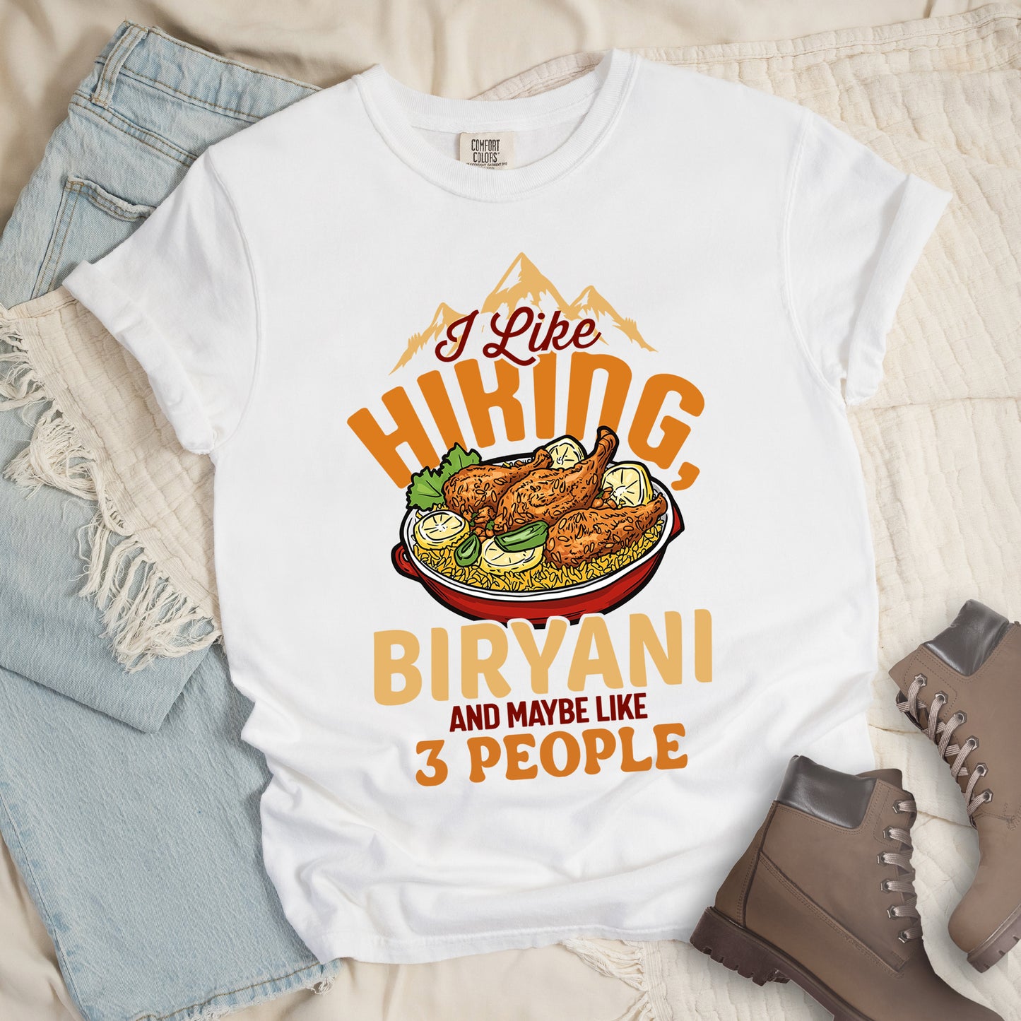 White t-shirt with text reading "I Like HIKING, BIRYANI AND MAYBE LIKE 3 PEOPLE". Gold mountain silhouettes appear above "I Like" (in dark brown script). "HIKING" is in large orange letters, below which sits a detailed illustration of a red bowl filled with biryani rice and chicken. "BIRYANI" appears in gold letters, while "AND MAYBE LIKE" is in small dark brown text and "3 PEOPLE" in orange. Features a classic crew neck and casual rolled sleeves, styled alongside light wash denim and brown hiking boots.