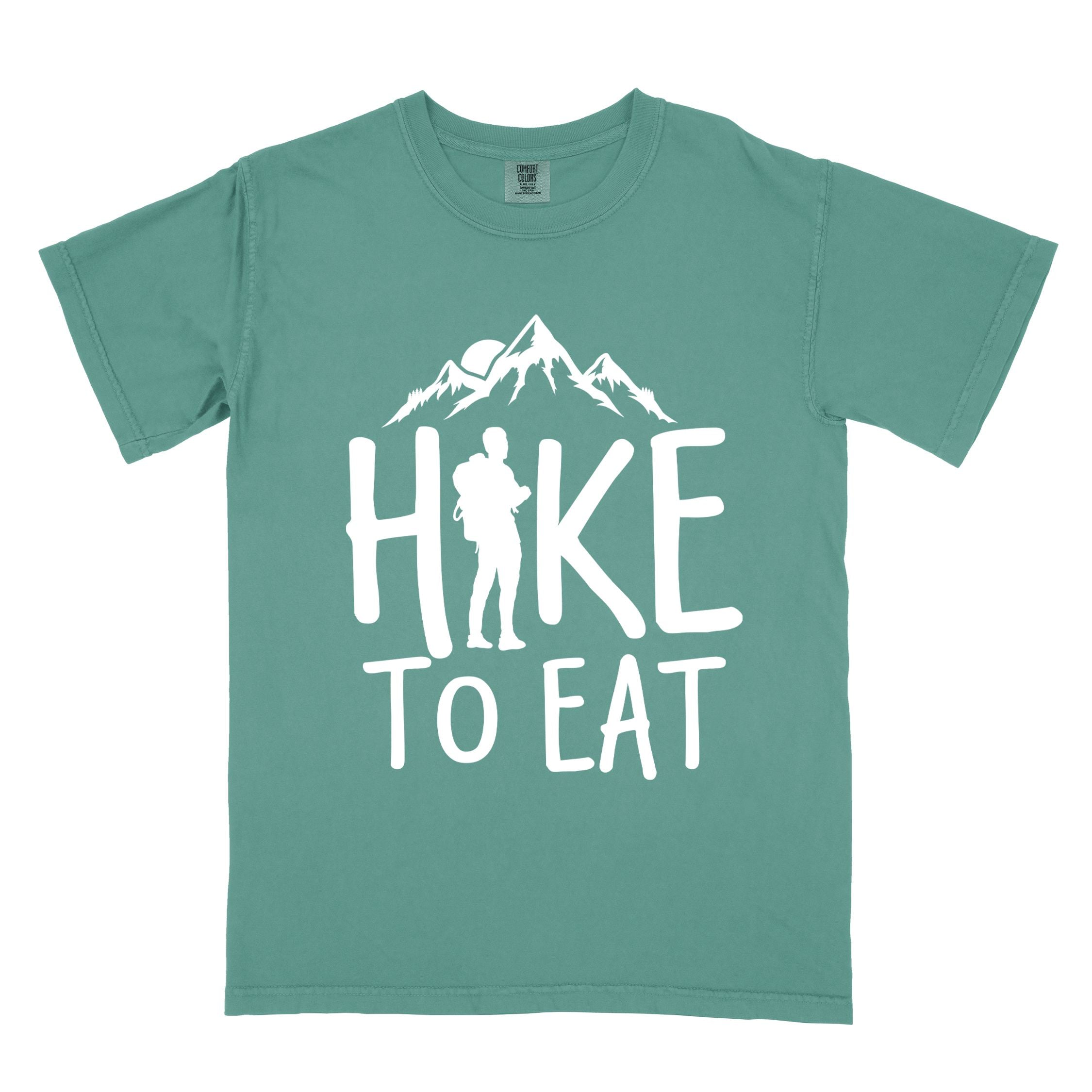 Light green "Hike to Eat" colored statement tee with bold typography, mountains, birds, sunrise, perfect for food motivated individuals.