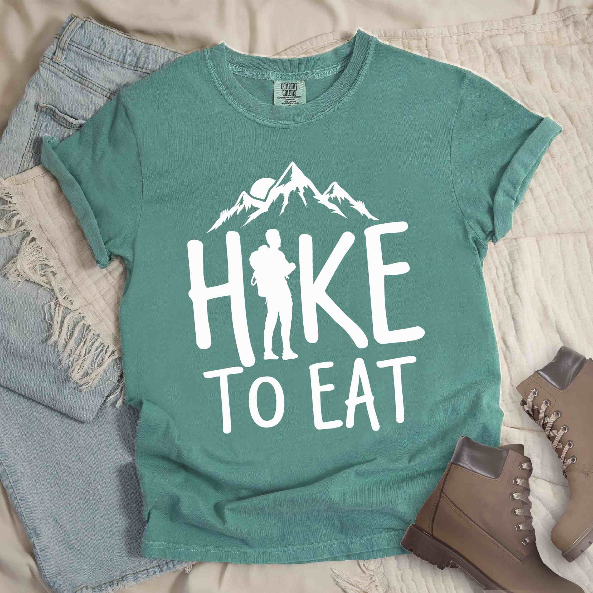 Light green "Hike to Eat" colored statement tee with bold typography, mountains, birds, sunrise, perfect for food motivated individuals.