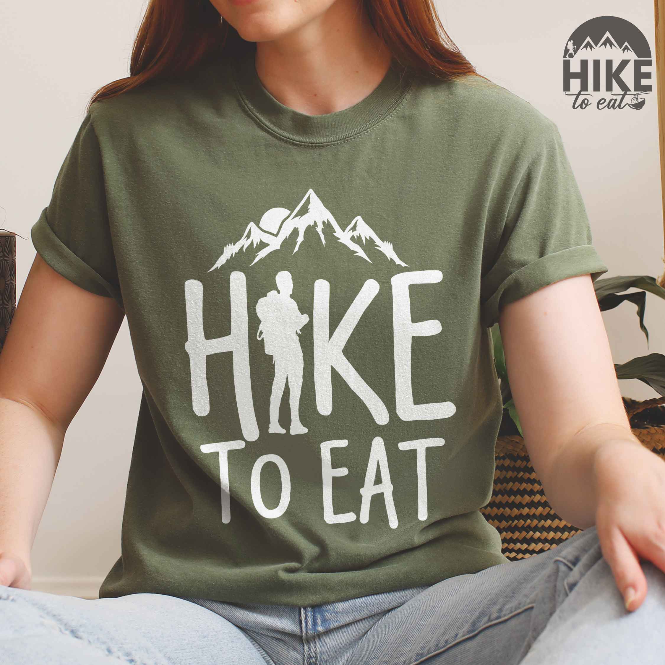 Woman wearing moss "Hike to Eat" colored statement tee with bold typography, mountains, birds, sunrise, perfect for food motivated individuals.