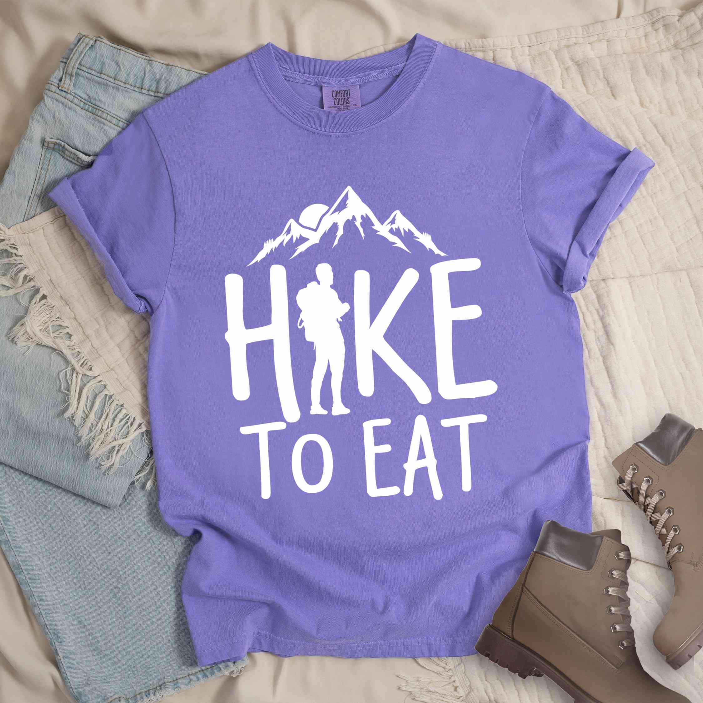 Violet "Hike to Eat" colored statement tee with bold typography, mountains, birds, sunrise, perfect for food motivated individuals.