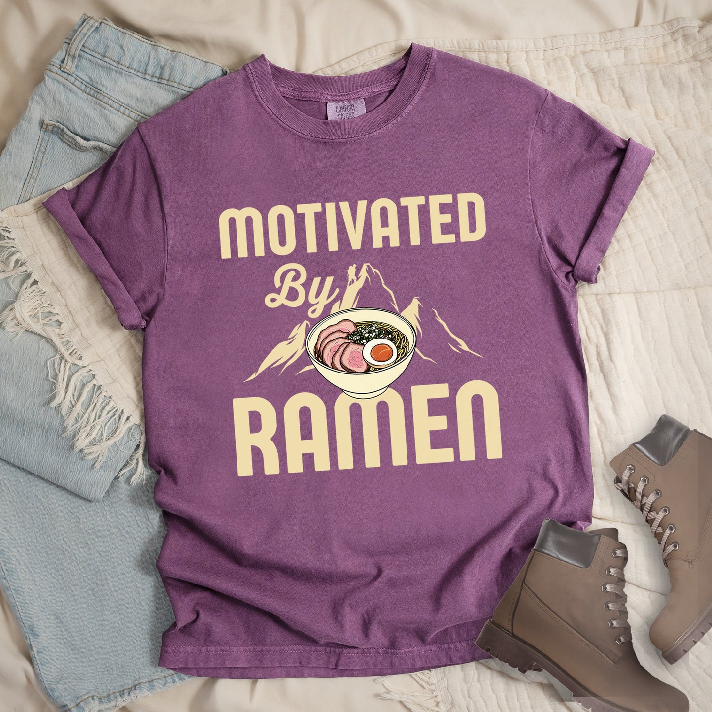 Berry (Dark Violet) t-shirt with cream-colored text reading "MOTIVATED By RAMEN". "MOTIVATED" appears in large letters at the top, "By" in script in the middle, and "RAMEN" in large letters at the bottom. Features a detailed illustration of a ramen bowl with sliced meat, egg, and green onions in the center, positioned over cream-colored mountain silhouettes with a small hiker figure on a peak.