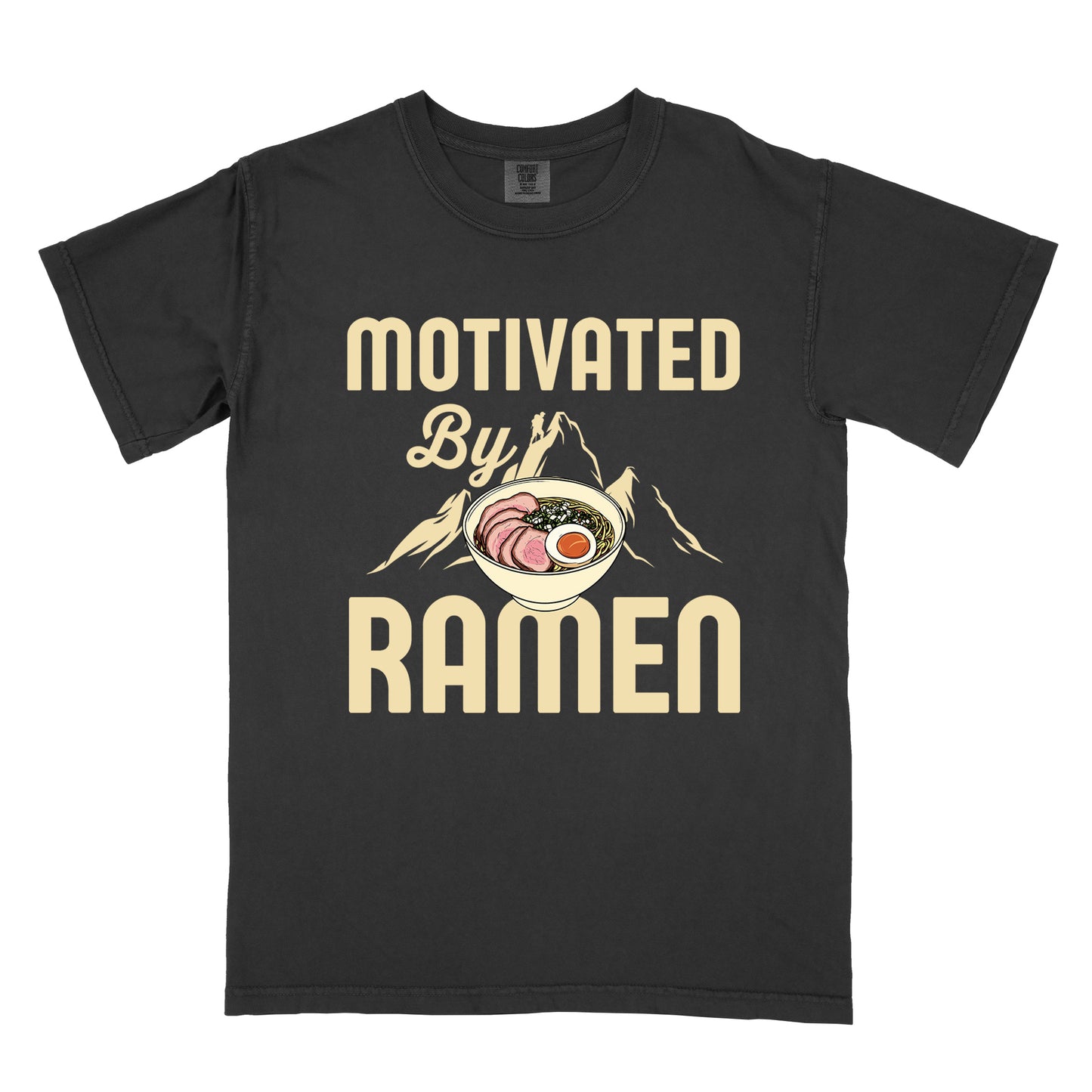 Black t-shirt with cream-colored text reading "MOTIVATED By RAMEN". "MOTIVATED" appears in large letters at the top, "By" in script in the middle, and "RAMEN" in large letters at the bottom. Features a detailed illustration of a ramen bowl with sliced meat, egg, and green onions in the center, positioned over cream-colored mountain silhouettes with a small hiker figure on a peak.