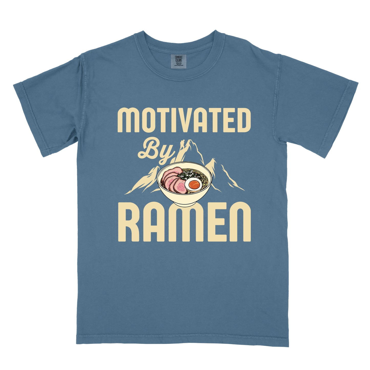 Blue Jean (Denim Blue) t-shirt with cream-colored text reading "MOTIVATED By RAMEN". "MOTIVATED" appears in large letters at the top, "By" in script in the middle, and "RAMEN" in large letters at the bottom. Features a detailed illustration of a ramen bowl with sliced meat, egg, and green onions in the center, positioned over cream-colored mountain silhouettes with a small hiker figure on a peak.