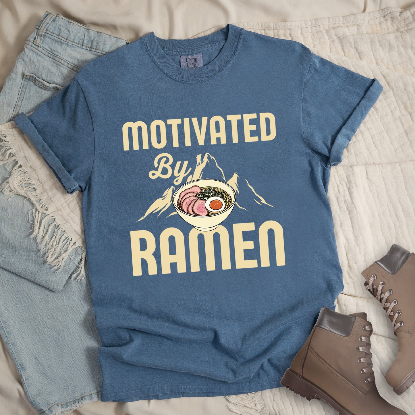 Blue Jean (Denim Blue) t-shirt with cream-colored text reading "MOTIVATED By RAMEN". "MOTIVATED" appears in large letters at the top, "By" in script in the middle, and "RAMEN" in large letters at the bottom. Features a detailed illustration of a ramen bowl with sliced meat, egg, and green onions in the center, positioned over cream-colored mountain silhouettes with a small hiker figure on a peak.