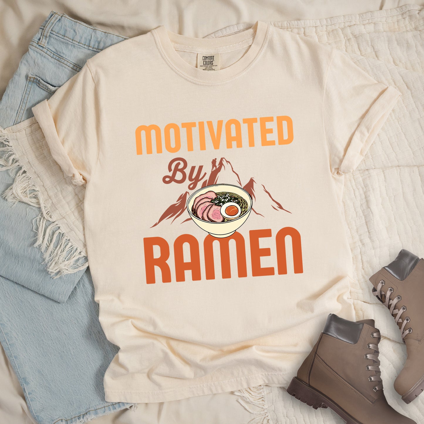 Ivory (Cream White) t-shirt with yellow, orange and brown-colored text reading "MOTIVATED By RAMEN". "MOTIVATED" appears in large letters at the top, "By" in script in the middle, and "RAMEN" in large letters at the bottom. Features a detailed illustration of a ramen bowl with sliced meat, egg, and green onions in the center, positioned over brown-colored mountain silhouettes with a small hiker figure on a peak.