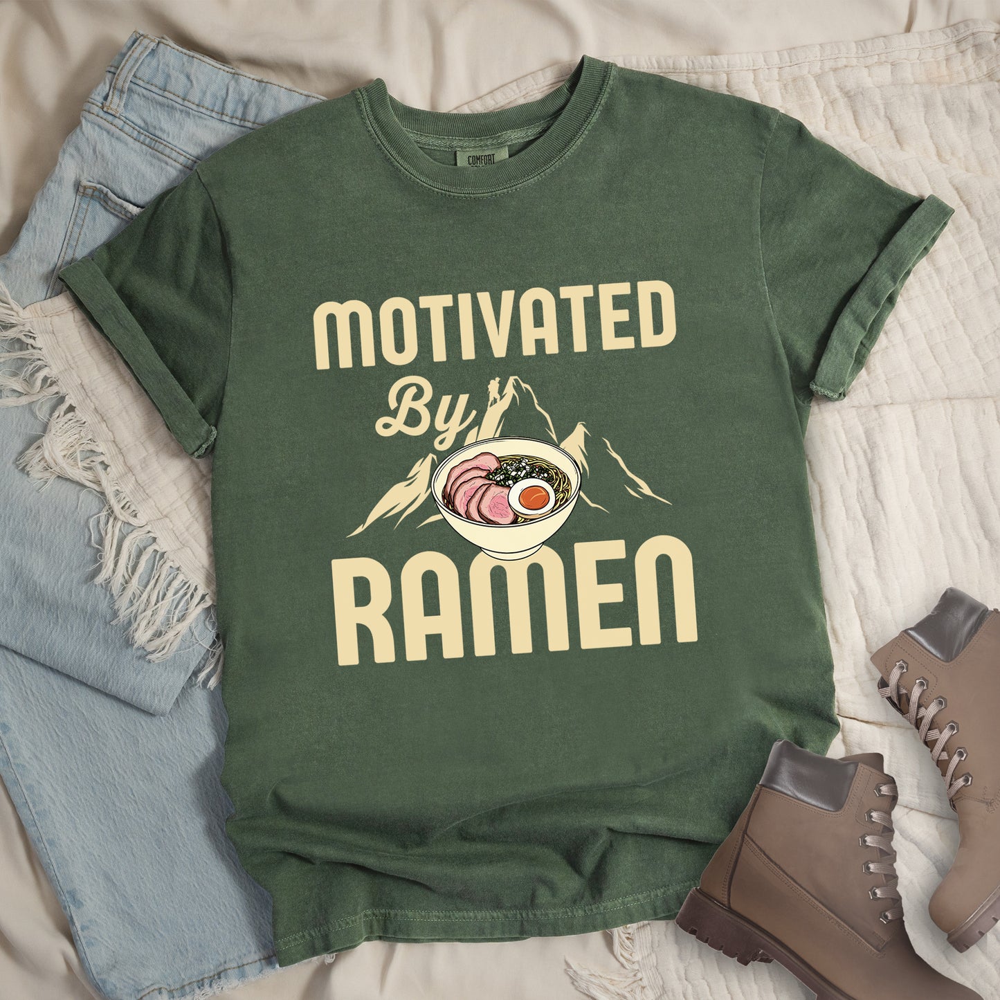 Moss (Dark Green) t-shirt with cream-colored text reading "MOTIVATED By RAMEN". "MOTIVATED" appears in large letters at the top, "By" in script in the middle, and "RAMEN" in large letters at the bottom. Features a detailed illustration of a ramen bowl with sliced meat, egg, and green onions in the center, positioned over cream-colored mountain silhouettes with a small hiker figure on a peak.