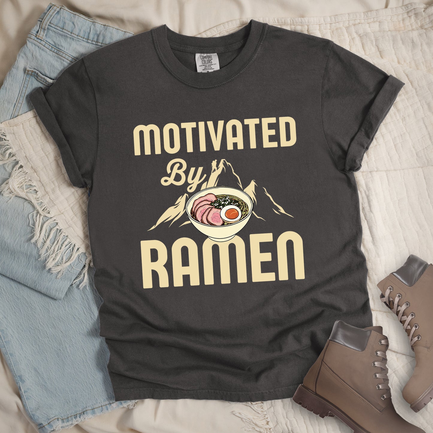 Pepper (Dark Gray) t-shirt with cream-colored text reading "MOTIVATED By RAMEN". "MOTIVATED" appears in large letters at the top, "By" in script in the middle, and "RAMEN" in large letters at the bottom. Features a detailed illustration of a ramen bowl with sliced meat, egg, and green onions in the center, positioned over cream-colored mountain silhouettes with a small hiker figure on a peak.