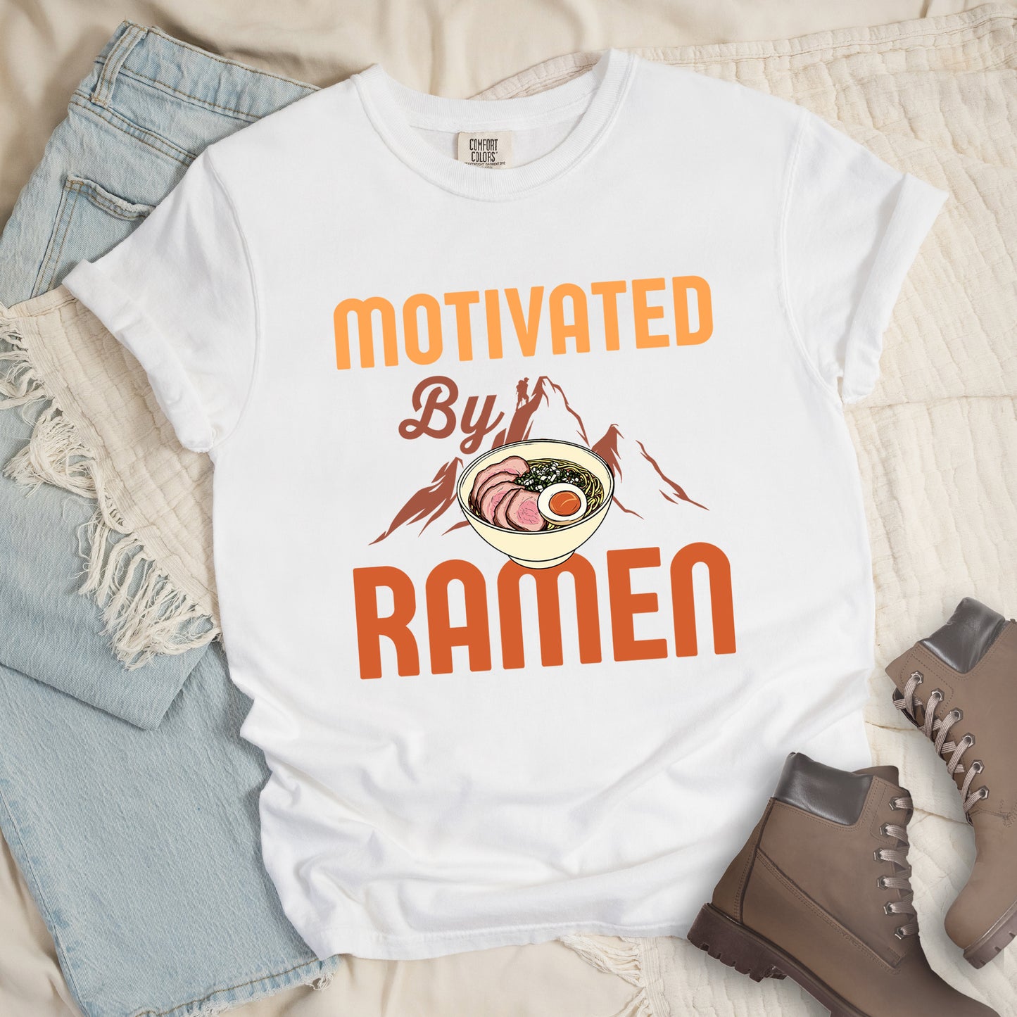 White t-shirt with yellow, orange and brown-colored text reading "MOTIVATED By RAMEN". "MOTIVATED" appears in large letters at the top, "By" in script in the middle, and "RAMEN" in large letters at the bottom. Features a detailed illustration of a ramen bowl with sliced meat, egg, and green onions in the center, positioned over brown-colored mountain silhouettes with a small hiker figure on a peak.
