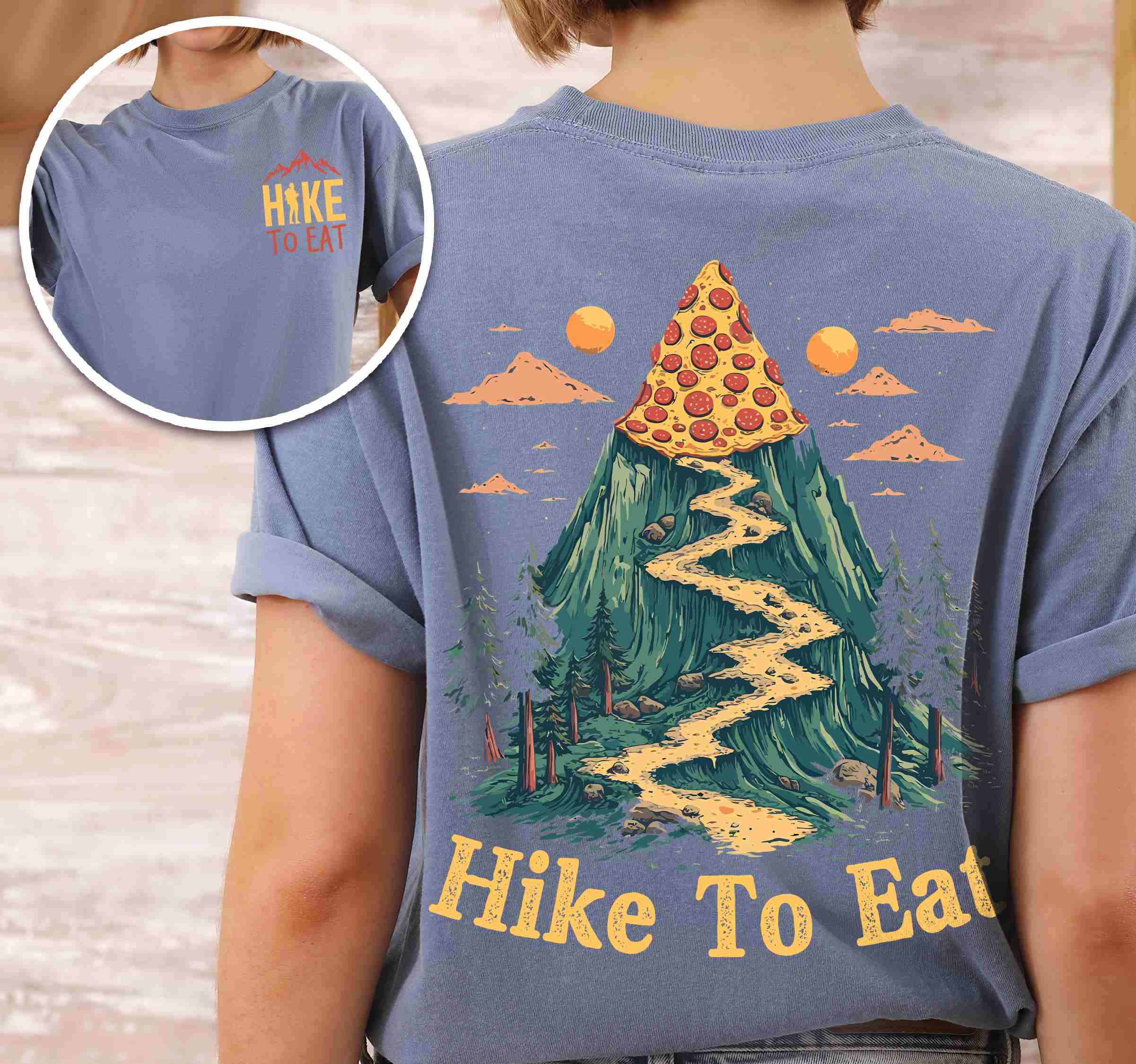 Woman wearing Blue jean colored vintage tee with front and back design "Hike to Eat" text in cream lettering, mountains, birds, illustrated pizza slice mountain peak, and backpacker for pizza lovers.