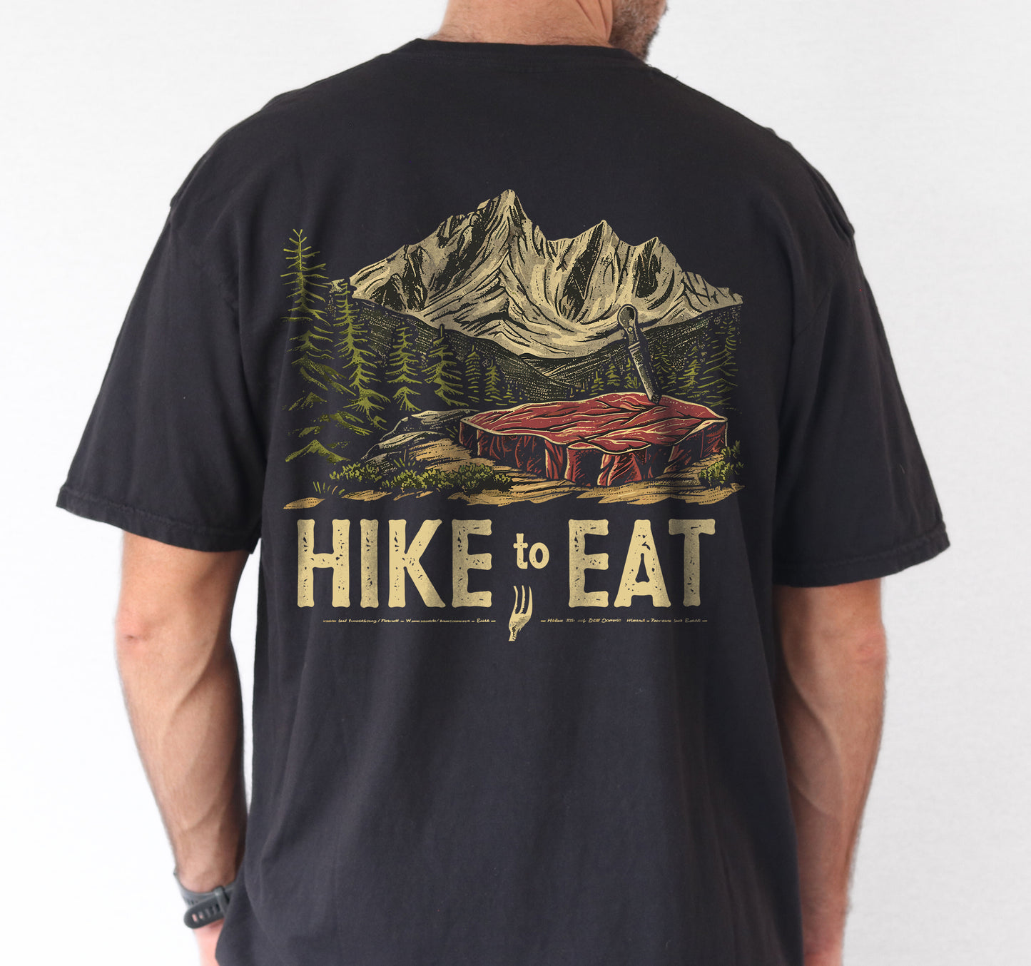 Black Hike to Eat t-shirt back view showing large design featuring raw steak in mountain wilderness landscape with pine trees worn by a male person.