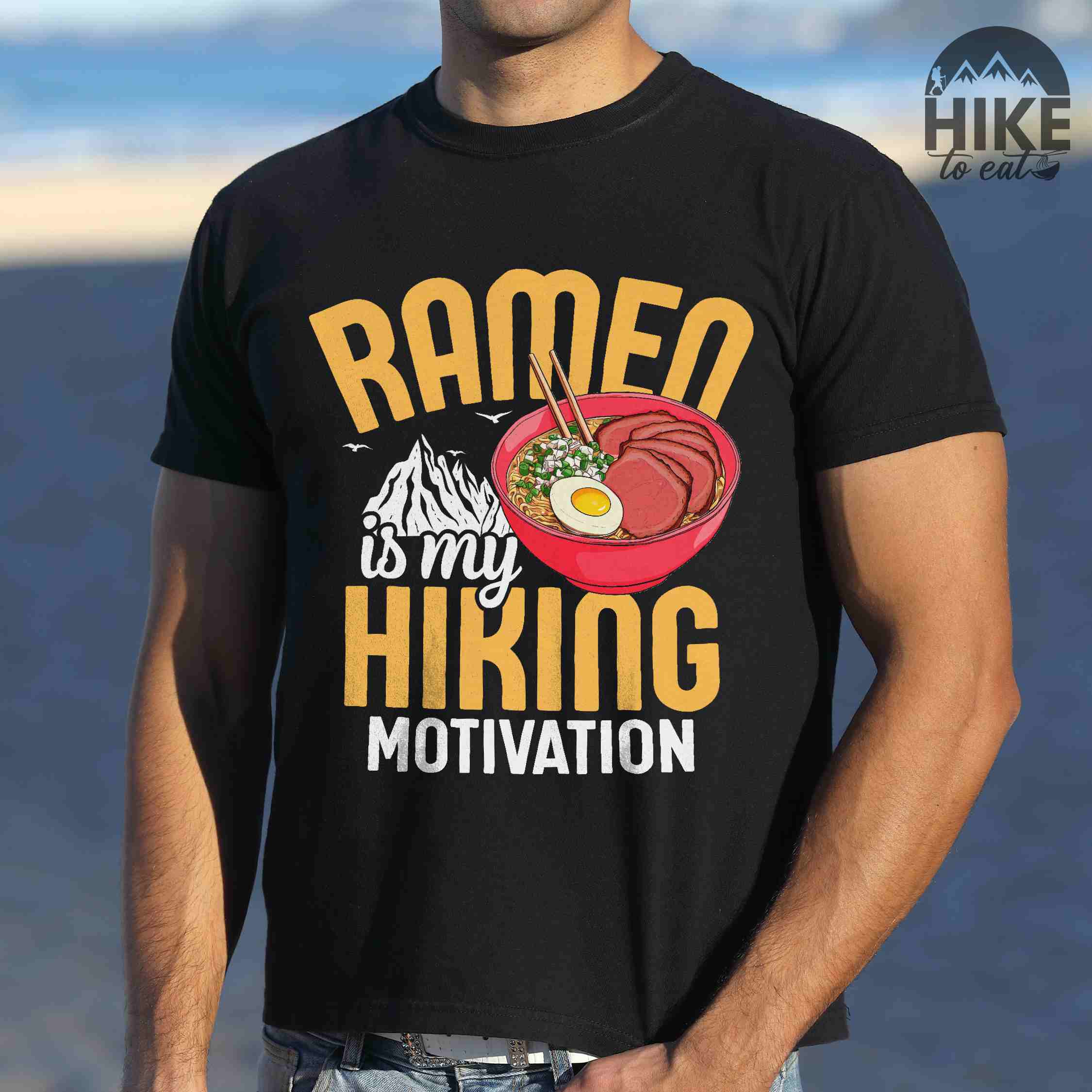 Man wearing black “Ramen is my Hiking motivation” colored vintage tee with bold text, mountain, Illustration of hot ramen. Perfect tee for foodie craving for a bowl of ramen,