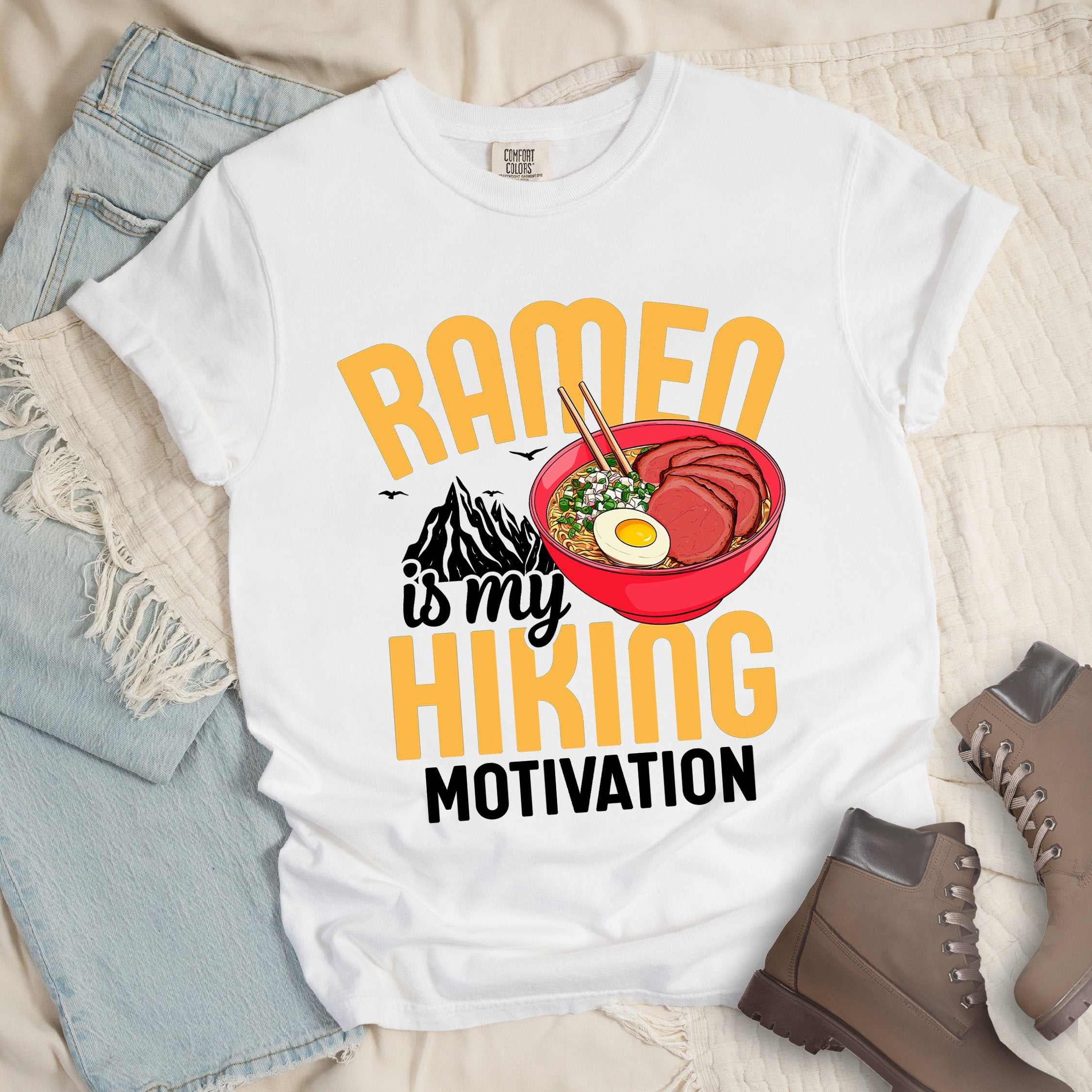 White shirt with “Ramen is my Hiking Motivation” text, featuring a mountain graphic and a ramen bowl with noodles, egg, meat, and chopsticks.