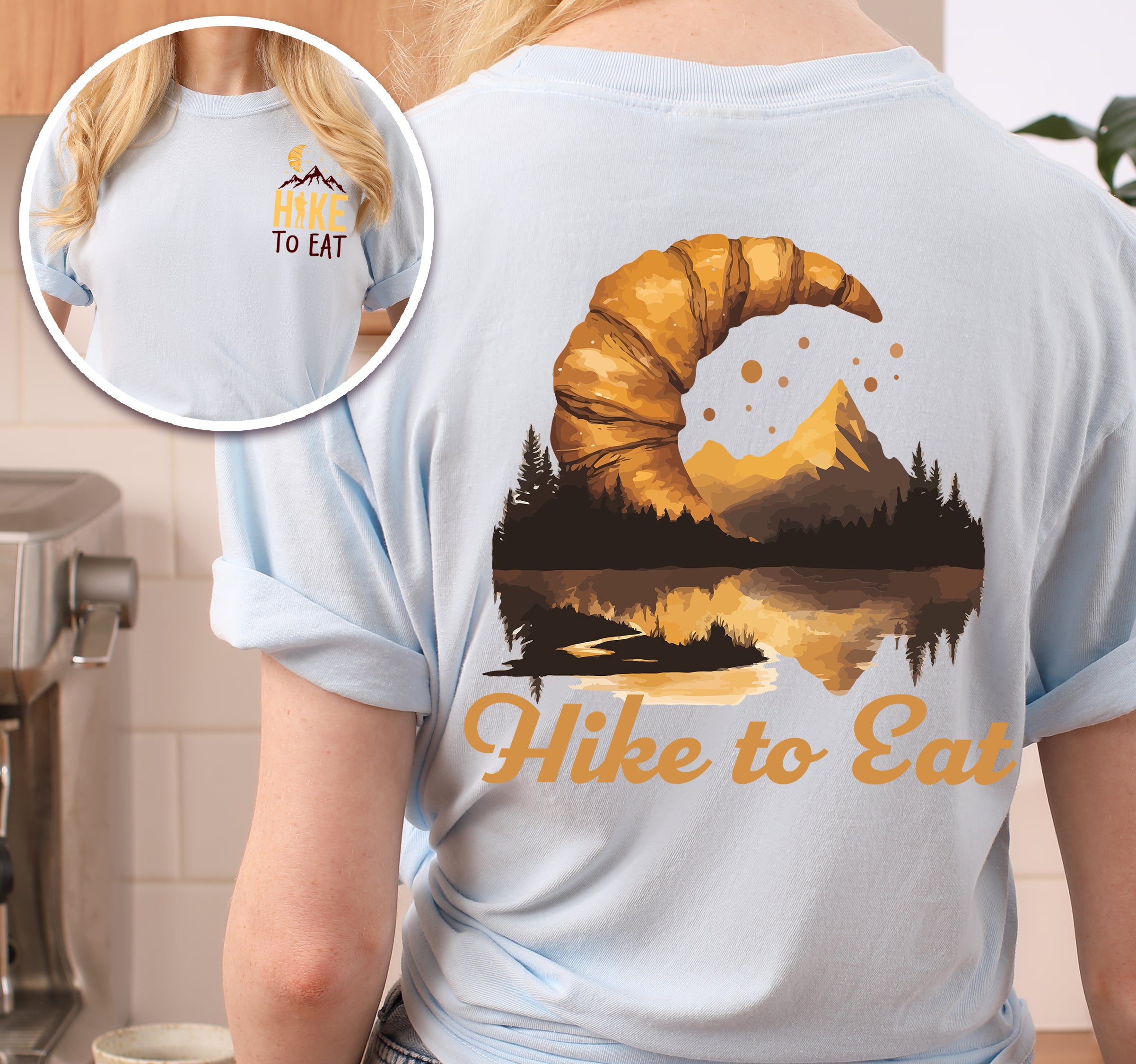 Chambray (Light Blue) Hike to Eat t-shirt front and back view showing small chest logo and large back design featuring golden croissant filled with sunset mountain lake landscape, pine trees, and reflected peaks.
