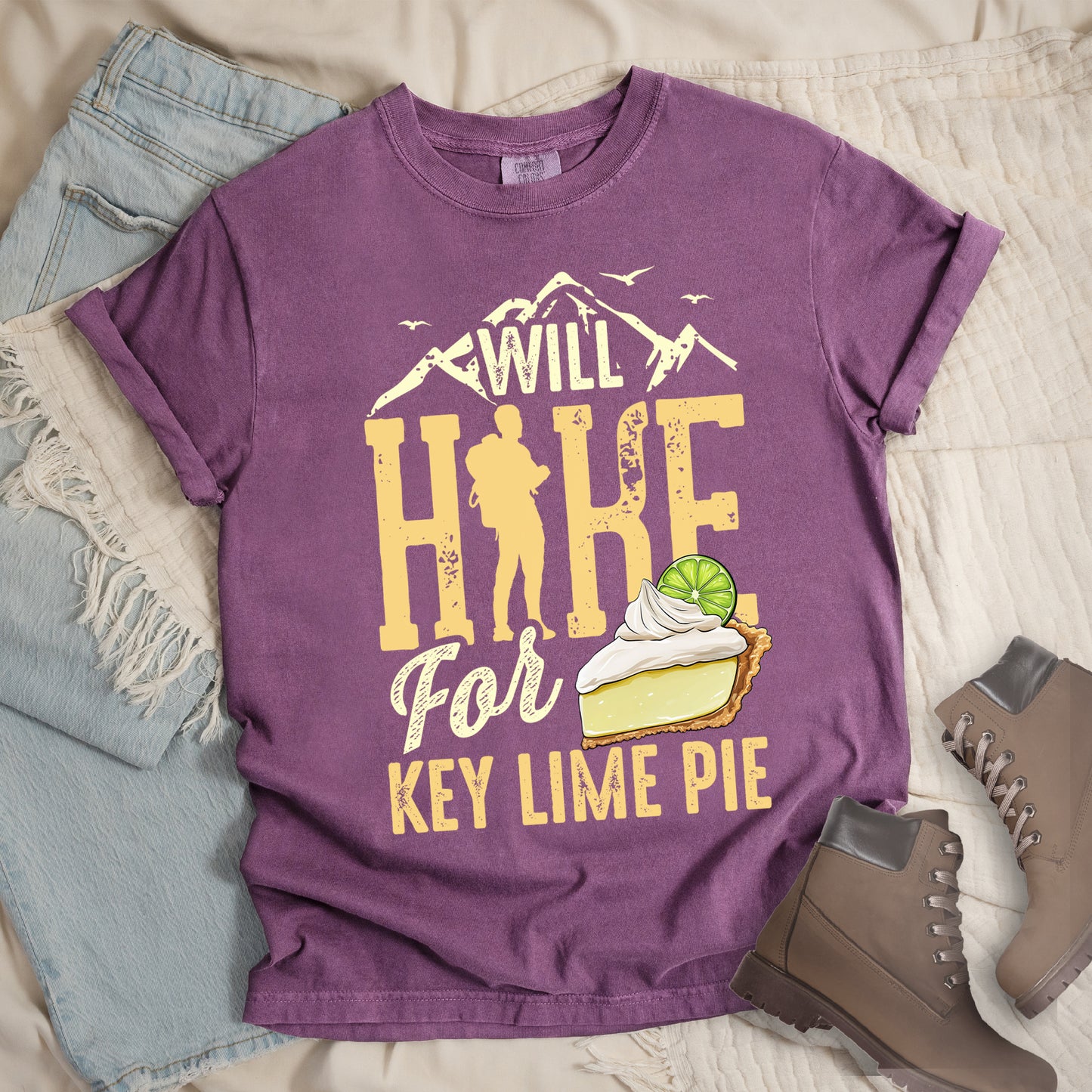 Berry (Dark Violet) t-shirt with text "WILL HIKE For KEY LIME PIE" in yellow tones. "WILL" appears in cream letters within mountain silhouettes with flying birds. "HIKE" is in large distressed yellow letters with a hiker silhouette replacing the "I". "For" appears in cream script, and "KEY LIME PIE" in yellow block letters. An illustration of a key lime pie slice with whipped cream and lime garnish appears next to "For" and above "KEY LIME PIE".