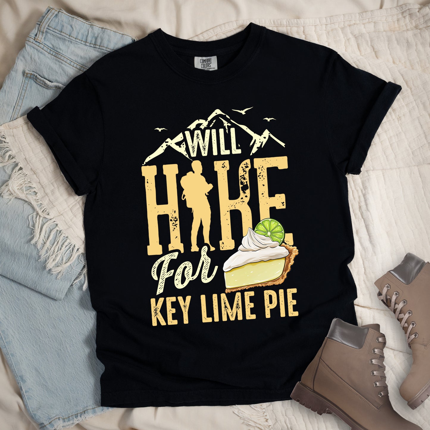 Black t-shirt with text "WILL HIKE For KEY LIME PIE." "WILL" appears in cream letters within mountain silhouettes with flying birds. "HIKE" is in large distressed yellow letters with a hiker silhouette replacing the "I". "For" appears in cream script, and "KEY LIME PIE" in yellow block letters. An illustration of a key lime pie slice with whipped cream and lime garnish appears next to "For." Features a classic crew neck and casual rolled sleeves, styled alongside light wash denim and brown hiking boots.