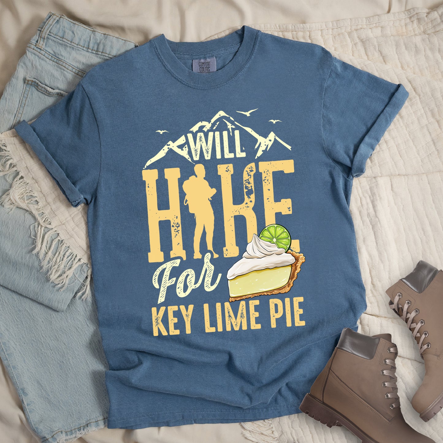 Blue Jean (Denim Blue) t-shirt with text "WILL HIKE For KEY LIME PIE" in yellow tones. "WILL" appears in cream letters within mountain silhouettes with flying birds. "HIKE" is in large distressed yellow letters with a hiker silhouette replacing the "I". "For" appears in cream script, and "KEY LIME PIE" in yellow block letters. An illustration of a key lime pie slice with whipped cream and lime garnish appears next to "For" and above "KEY LIME PIE".