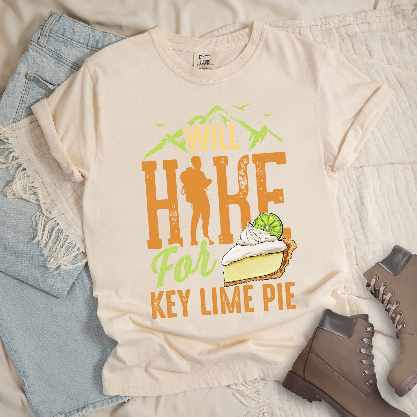 Ivory (Cream White) t-shirt with text "WILL HIKE For KEY LIME PIE" in orange and green tones. "WILL" appears in cream letters within mountain silhouettes with flying birds. "HIKE" is in large distressed orange letters with a hiker silhouette replacing the "I". "For" appears in light green script, and "KEY LIME PIE" in orange block letters. An illustration of a key lime pie slice with whipped cream and lime garnish appears next to "For" and above "KEY LIME PIE".