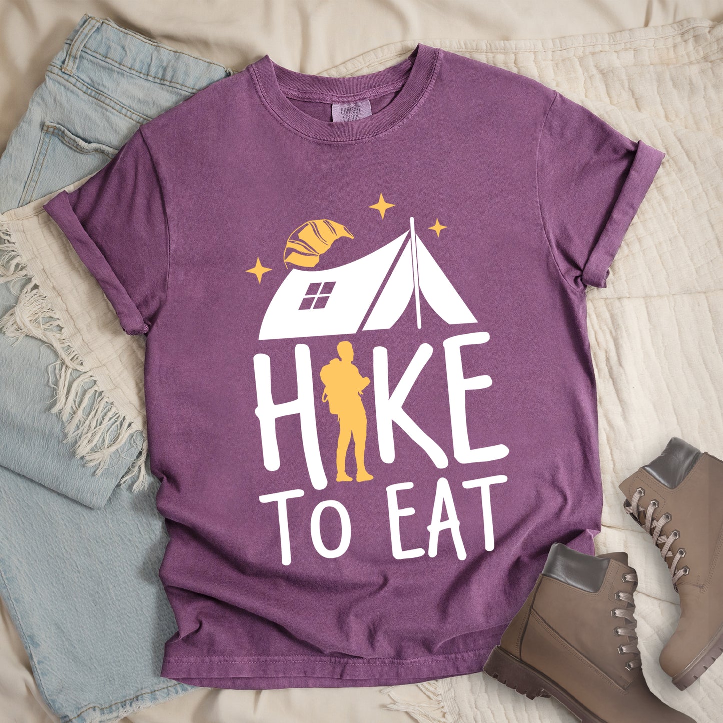 Berry (Dark Violet) graphic t-shirt with 'Hike to Eat' text featuring white camping tent, yellow hiker silhouette, croissant, and stars design, styled with light wash jeans and brown hiking boots.