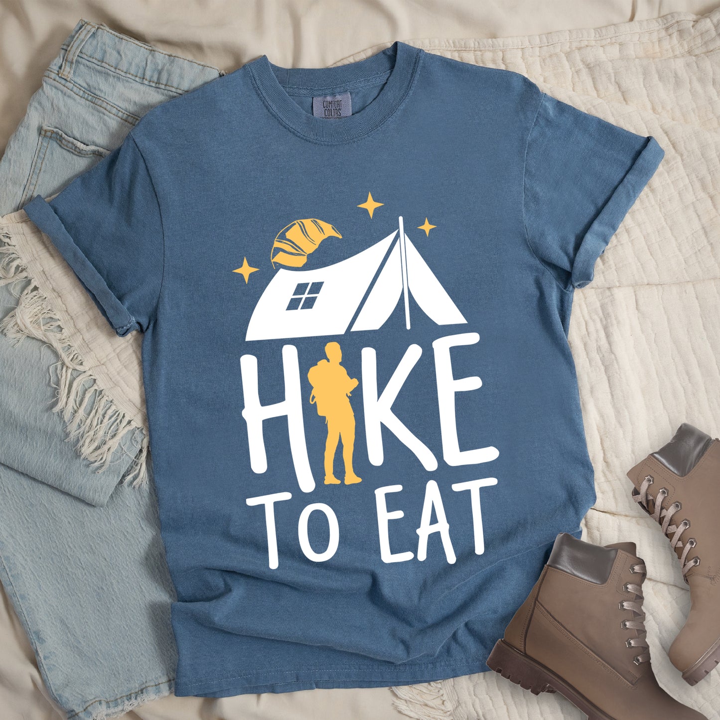 Blue Jean (Denim Blue) graphic t-shirt with 'Hike to Eat' text featuring white camping tent, yellow hiker silhouette, croissant, and stars design, styled with light wash jeans and brown hiking boots.